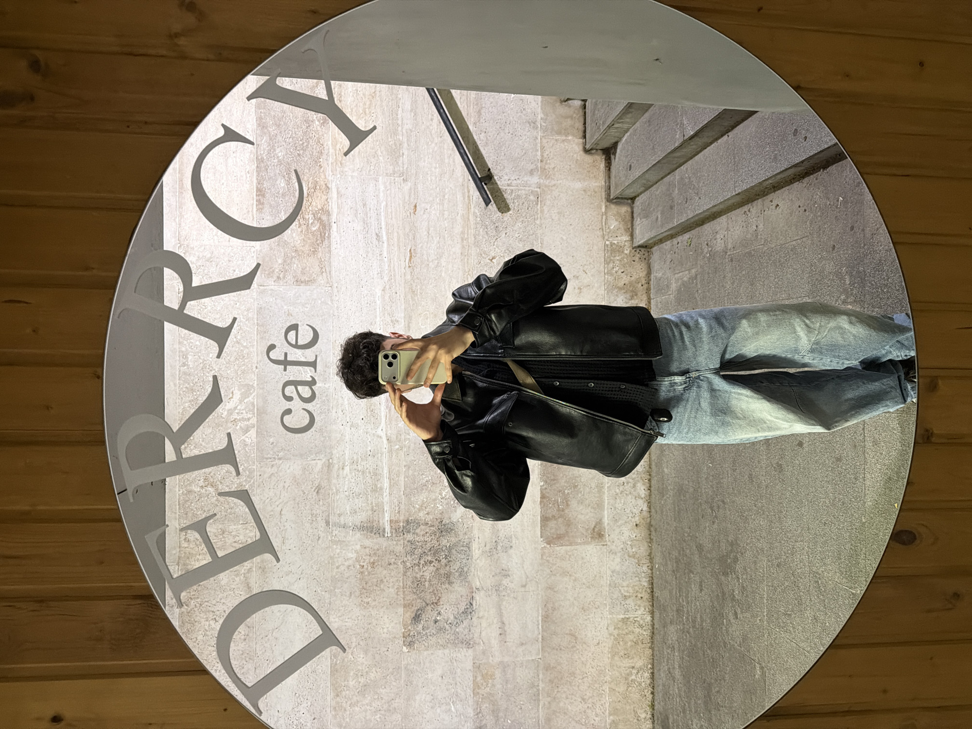 A person takes a selfie in a round mirror. The mirror's frame features a light gray inscription "D ERCY cafe." The reflection shows the person wearing a black leather jacket, a black textured top, and light blue jeans. They hold a smartphone in their right hand. The reflection also shows a stone wall behind them and a staircase. The mirror is placed against a wall with light brown wooden paneling. Based on the cafe name, the location is likely in Tbilisi, Georgia, due to the "Dercy" brand presence there.