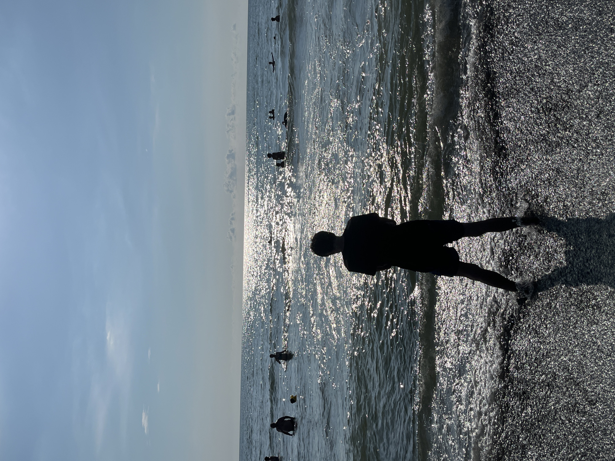 A person stands on a dark pebble beach, facing the sea. The sun glints off the water, creating a shimmering effect. Several other people are visible in the water, some swimming and others standing. The sky is a clear blue with a few scattered clouds. The location is Ureki, Georgia, a town known for its magnetic black sand beaches.
