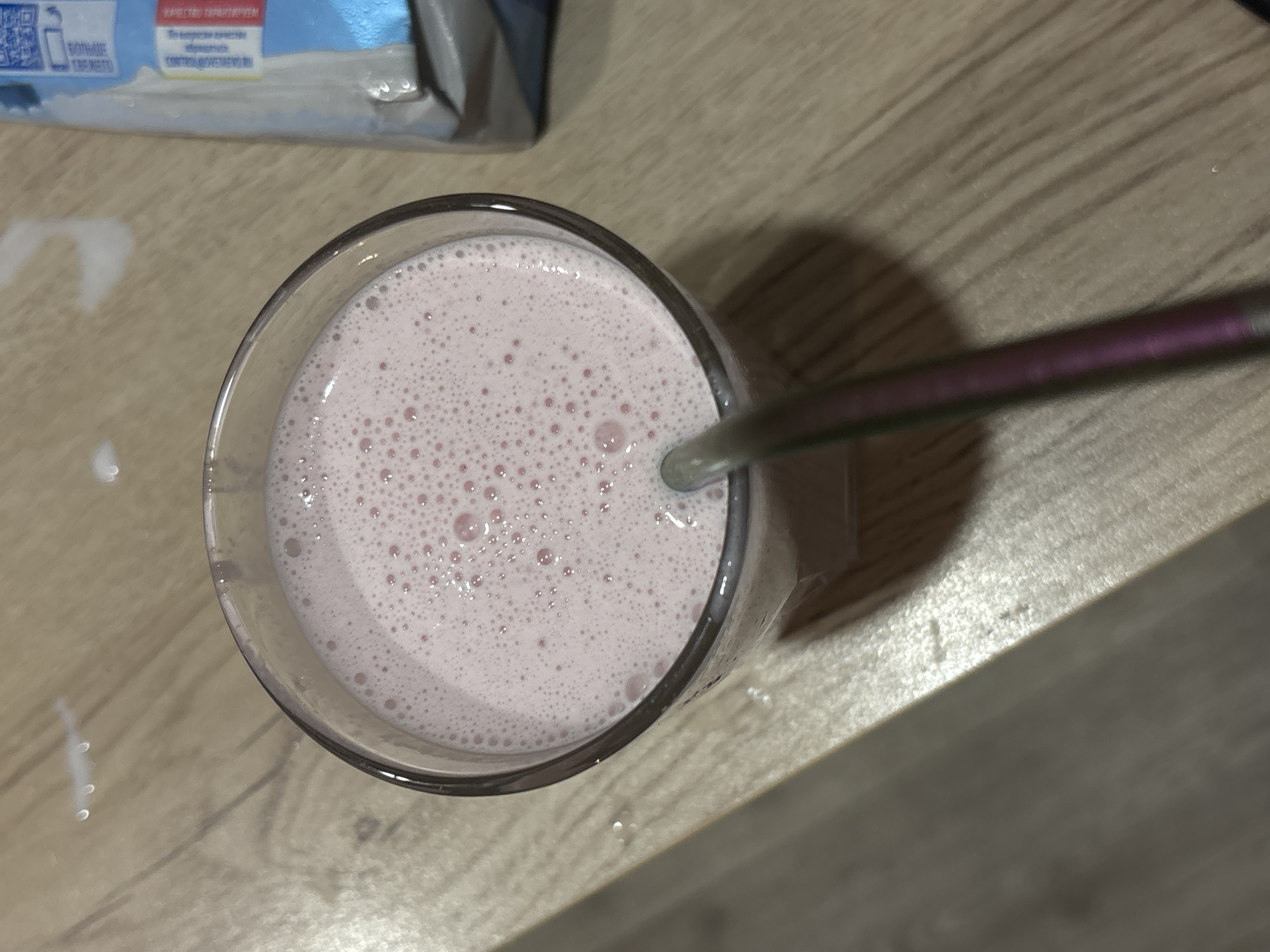 An overhead shot shows a glass filled with a light pink, bubbly liquid. A silver-purple straw is partially submerged in the drink. A package is slightly visible in the upper left corner, with text written in Russian. The surface underneath appears to be a light wood-grain laminate. The location is Chaikovsky, Russia.
