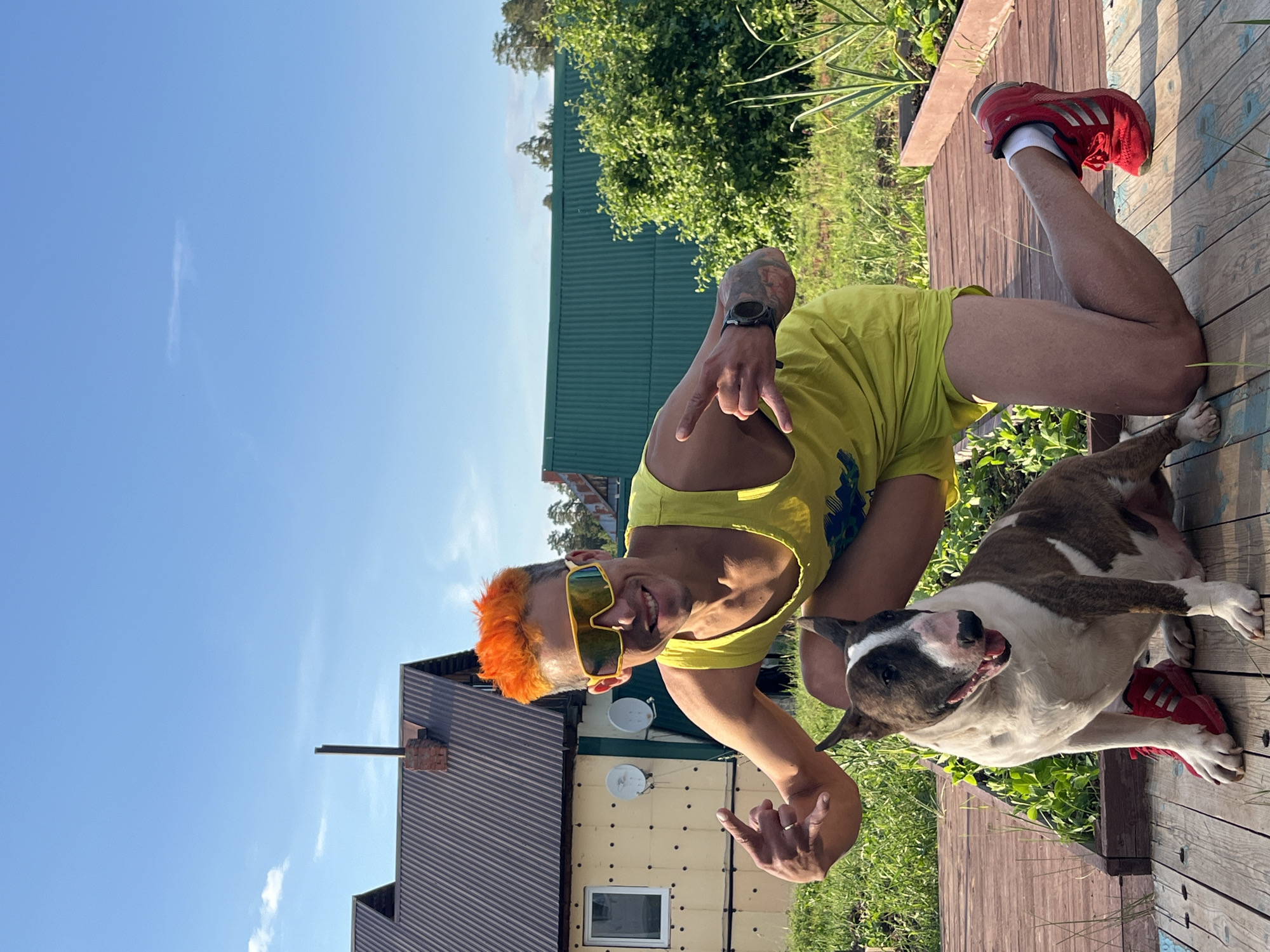 A man with bright orange hair and yellow sunglasses is kneeling on a wooden deck next to a brindle and white bull terrier. The man is wearing a yellow tank top and yellow shorts, and a black watch on his left wrist. He is making a rock-and-roll hand gesture with his right hand and a peace sign with his left. The dog is wearing a red sneaker on its left front paw. In the background are two houses with dark metal roofs, one light yellow and one green. There is also a large green bush and a blue sky with a few clouds. The scene appears to be in a rural or suburban area, possibly in Chaikovsky, Russia, given the style of the houses and the vegetation.