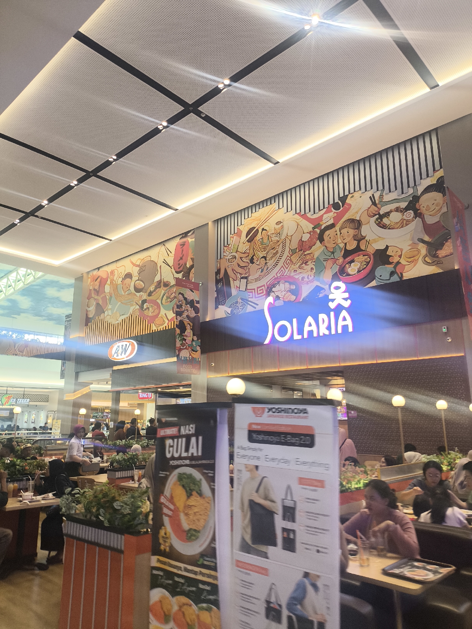 The image shows the interior of a food court in Grogol, Indonesia. In the foreground, there is a large banner advertising "Yoshinoya Japanese Restaurant" and their "Yoshinoya E-Bag 2.0". The banner features images of bags and descriptions like "Everyone Everyday Everything". To the left of the banner, there is another sign for "Nasi Gulai". People are seated at tables in the background, some eating and some talking.
Above the food court area, a large mural depicting cartoon characters and food is painted on the wall. The mural is bright and colorful, featuring scenes of people eating and preparing food. The name "SOLARIA" is prominently displayed in large, neon-like letters beneath the mural, illuminated in blue. A sign for "AW" (A&W Restaurants) is also visible on the left side of the mural. The ceiling is modern with a grid-like structure and integrated lighting. The overall impression is a busy and well-lit dining area within a mall or similar establishment.