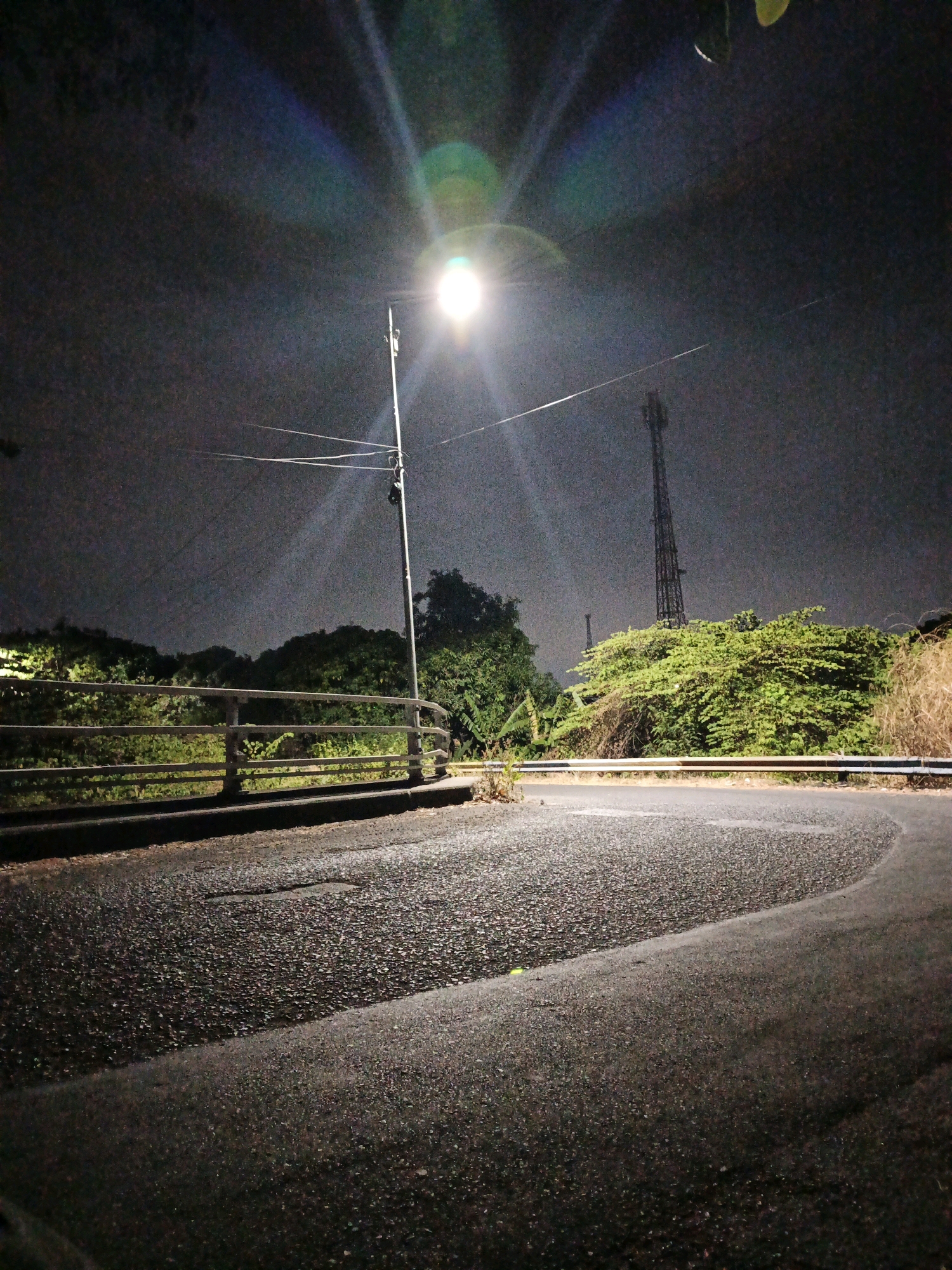 A low-angle shot depicts a paved road at night, curving to the right and away from the viewer. The road surface is asphalt with visible pebbles. A metal guardrail runs along the left side of the road, separating it from a vegetated area. In the background, lush green trees and bushes are illuminated by a bright streetlamp on the right side of the image. The streetlamp emits a strong light that creates lens flares in the dark sky. Further in the background, a tall, lattice-like telecommunications tower is visible. The scene appears to be in Kludan, Indonesia, likely a rural or semi-rural area on the outskirts of a city, given the presence of the tower and the vegetation. There are no people or animals visible in the image.
