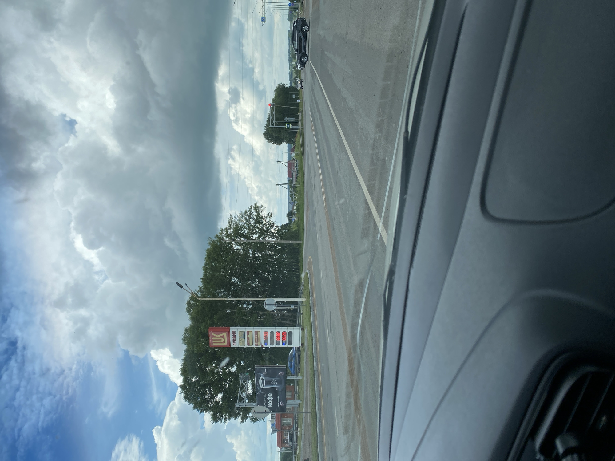 The image shows a view from inside a vehicle, looking out at a roadside scene in Kushnarenkovo, Russia. A Lukoil gas station sign with prices for various fuels is prominently displayed on the right. A billboard advertising coffee is visible on the left. The road ahead is wet, suggesting recent rain, and a few cars are present on the road. Trees line the background, and the sky is filled with dramatic clouds. The scene appears to be a typical view of a gas station and intersection in a rural or suburban area.