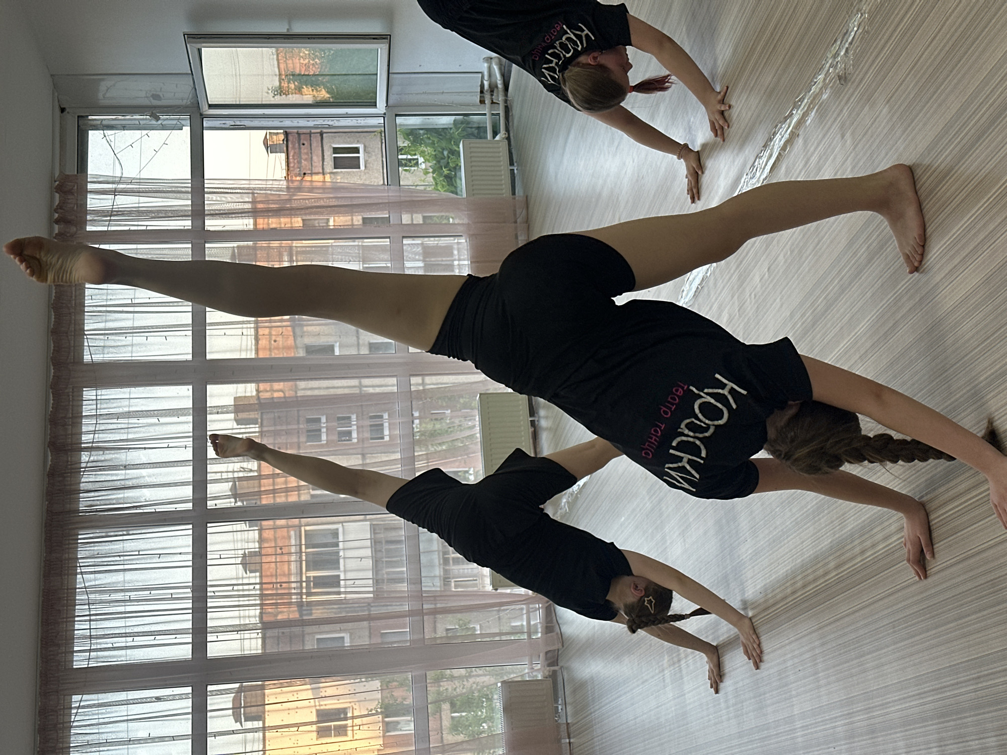 The image shows three young females, likely dancers, in a dance studio in Votkinsk, Russia. They are wearing black t-shirts with the word "КРАСКИ" (Paints) and the phrase "театр танца" (dance theatre) written on them. The studio has large windows, through which buildings in Votkinsk are visible. The women are performing acrobatic stretches, with their legs extended high. The background includes the studio floor, windows, and exterior buildings.