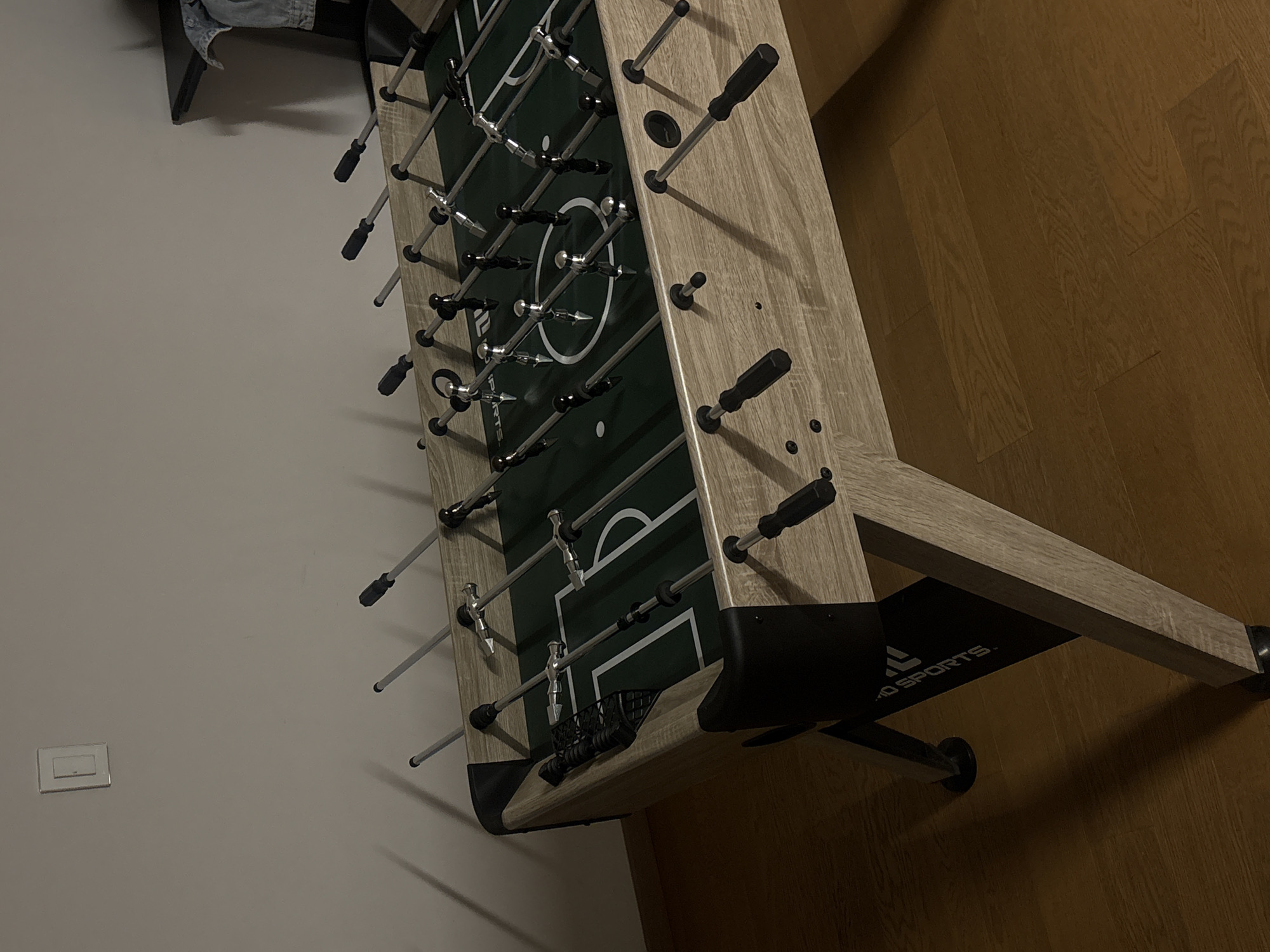 A slightly angled, high-angle indoor shot depicts a foosball table situated near a wall with an electrical outlet and a wooden floor. The table is primarily light wood with black and silver components. Visible text on the table reads "Sports". The room's lighting is dim. The location is unspecified and there is no way to know if this image was taken in San Bernardino Tlaxcalancingo, Mexico.