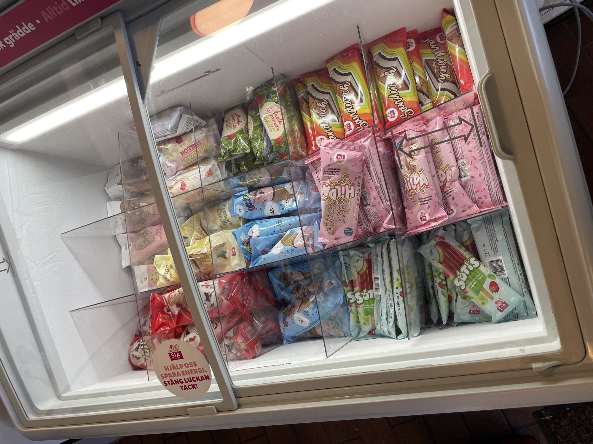 The image shows the interior of an open freezer display case, likely in a convenience store or supermarket, filled with a variety of packaged ice cream and frozen treats. The freezer is organized into several clear plastic compartments, each holding different types of frozen desserts.
In the upper left section, there are several red and white packaged ice creams, and a circular red and white sticker with Swedish text is visible. Moving to the right, various ice cream bars and cones are displayed in their packaging, some with colorful graphics and text in Swedish, such as "Hallon" (raspberry) and "Lemon Curd."
In the middle section, there are ice cream bars with blue packaging featuring cartoon characters, and more colorful ice cream bars with various flavors. Further to the right, several individually wrapped ice cream sandwiches with the brand name "Sandwich" are stacked.
The bottom section reveals more ice cream bars, some with green and white packaging indicating flavors like apple, and others with pink packaging featuring sprinkles. A bright pink ice cream bar is partially visible in the lower right corner.
The freezer unit itself appears to be white and is well-lit by internal lighting, which causes some glare and frost on the surfaces. The overall impression is a well-stocked freezer ready for customers.
The presence of Swedish text on the packaging and the specific mention of Karlstad, Sweden, in the prompt suggests this freezer is located in a retail establishment within that city. Without further context or visible landmarks, the precise location within Karlstad cannot be determined from the image alone. The scene is devoid of people or any actions being performed.