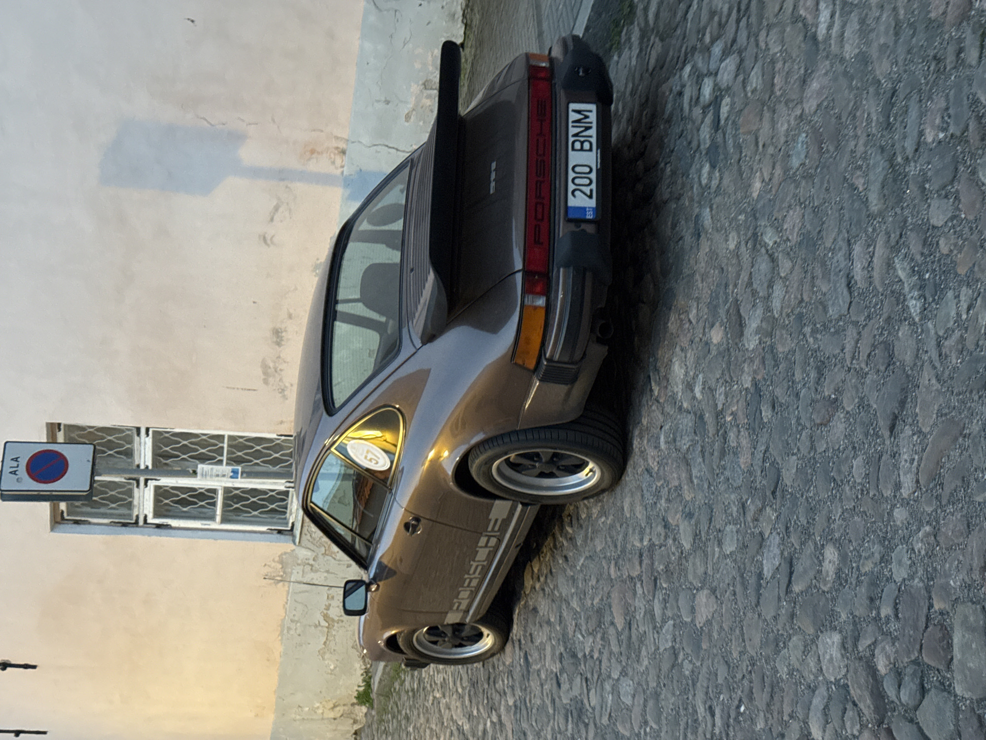 A brown Porsche 911 is parked on a cobblestone street in Tallinn, Estonia. The car is facing away from the viewer, showing its rear and right side. It has a black rear spoiler and a license plate that reads "200 BNM". The car is parked in front of a building with a cream-colored facade and a window with a patterned grate. A street sign with the word "ALA" and a parking symbol is visible above the window. The lighting suggests it is either early morning or late afternoon. The cobblestone street is uneven and appears old. There are no people visible in the image.