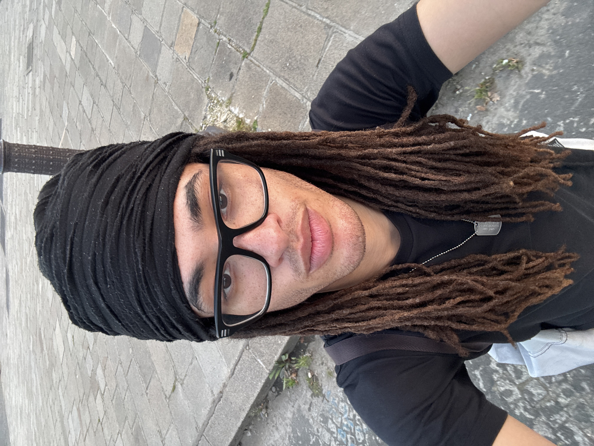 A selfie from a high angle shows a man with dreadlocks and glasses, partially covered by a black head wrap. The man is wearing a black t-shirt and a silver dog tag necklace. He is standing on a paved street, possibly a sidewalk, consisting of rectangular grey stones laid in a running bond pattern. The bottom left corner features a dark, cylindrical object, possibly a pole or bollard. Patches of greenery are visible between the paving stones. Given the architectural style and street layout, the location is likely within the city of Paris, France.