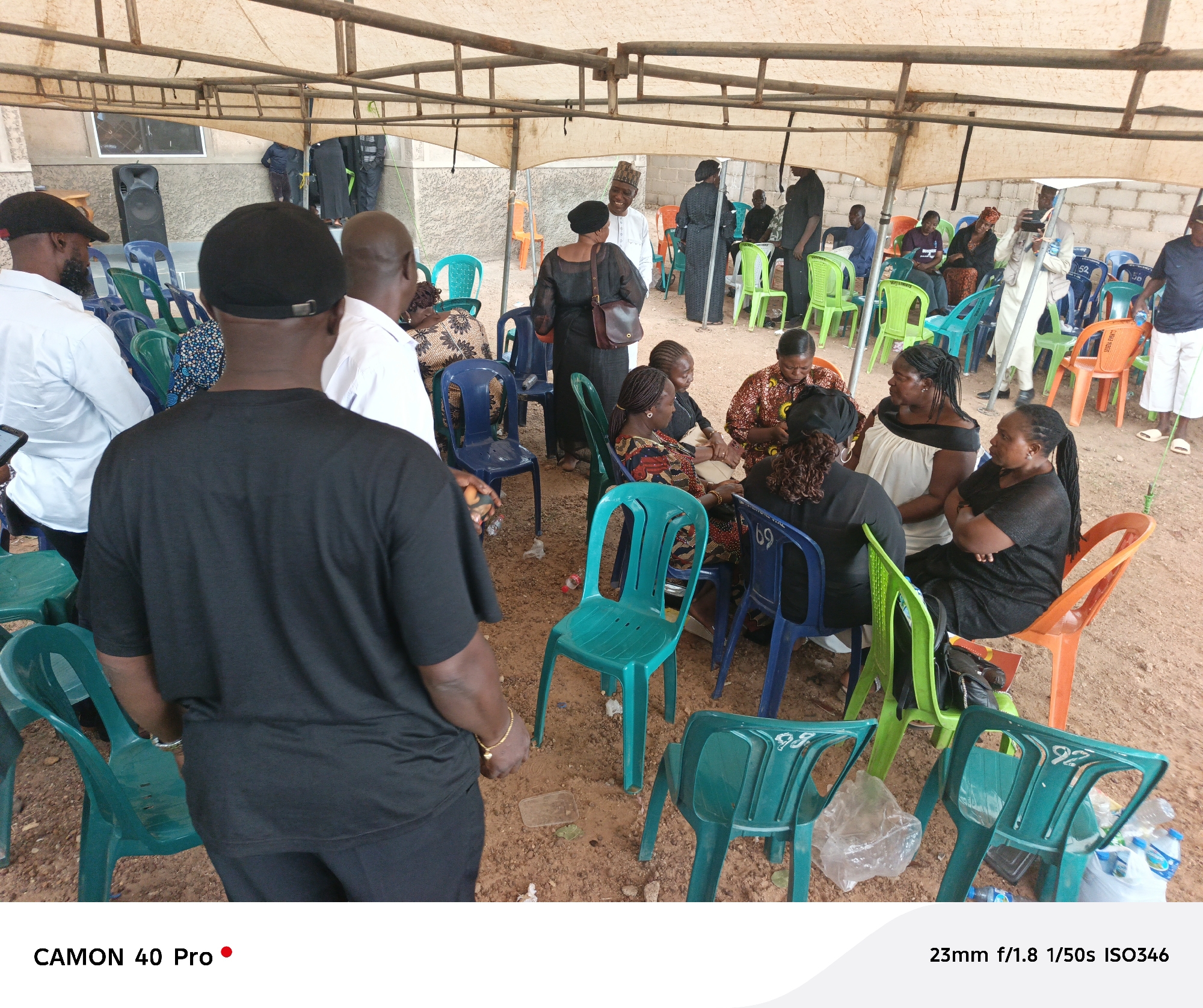 The image captures a gathering under a canopy in what appears to be an outdoor event in Jos, Nigeria. The ground is dirt. The canopy is supported by a metal frame. A mix of people are present, seated on a variety of plastic chairs in green, blue, and orange colors. A group of women are seated together, some wearing black or patterned clothing. Some men stand, wearing white shirts. In the background, more individuals are standing or sitting, some dressed in traditional attire. Water bottles and debris are visible on the ground around the chairs. In the distance, there is a building with what looks like a speaker outside it.