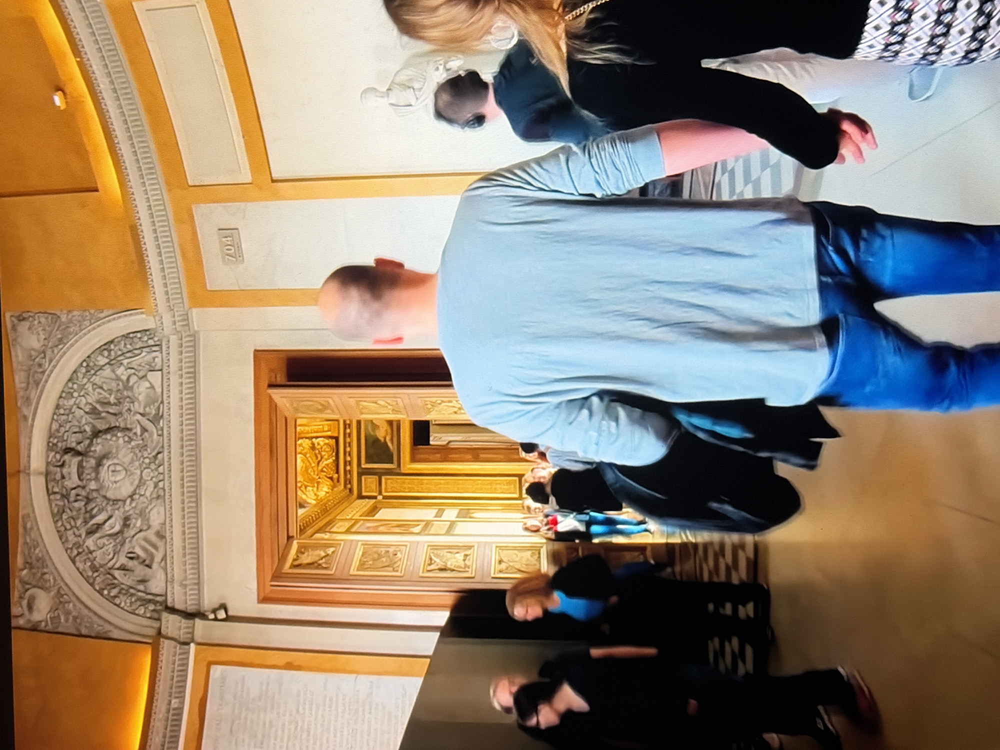 The image depicts an interior scene of what appears to be a museum or art gallery. In the foreground, a bald man wearing a light blue long-sleeved shirt and blue jeans is walking away from the camera. To his right, a woman with blonde hair is holding his hand. She is wearing a black long-sleeved top and patterned pants. Behind them, other museum visitors are visible, some walking and some standing. The environment features ornate architecture, including a large arched doorway leading to a richly decorated room, columns, and detailed moldings. A sign with the number "704" is visible on a wall. The lighting is warm, suggesting artificial lighting within the building. The floor is a light-colored tiled pattern in some areas and a smooth, polished surface in others.