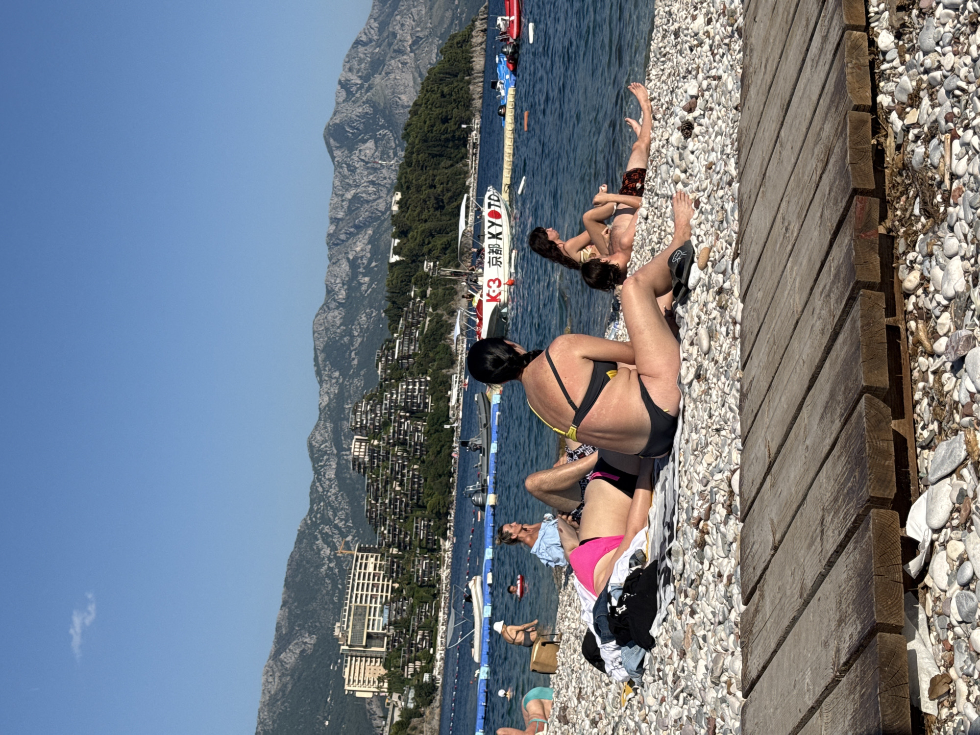 The image shows a sunny day at a pebble beach in Budva, Montenegro. Several people are relaxing on the beach, some lying on towels and others sitting up. The water is a deep blue and appears calm, with a few small boats and a yellow inflatable ring visible in the distance.
In the foreground, a wooden walkway runs along the edge of the beach. The people on the beach are dressed in swimwear. One woman in a yellow and black bikini is sitting up, facing away from the camera, while another woman in a pink bikini lies beside her. A man in patterned shorts is also visible, lying down further up the beach.
In the background, a white boat with red lettering that appears to say "K3 京都市 KYOTO" is moored near the shore. Beyond the beach, lush green trees cover the slopes of steep, rocky mountains, dotted with numerous buildings that form a dense urban landscape. The overall scene suggests a popular tourist destination during warm weather, likely mid-day given the strong sunlight casting shadows. The atmosphere appears relaxed and leisurely, typical of a vacation setting.