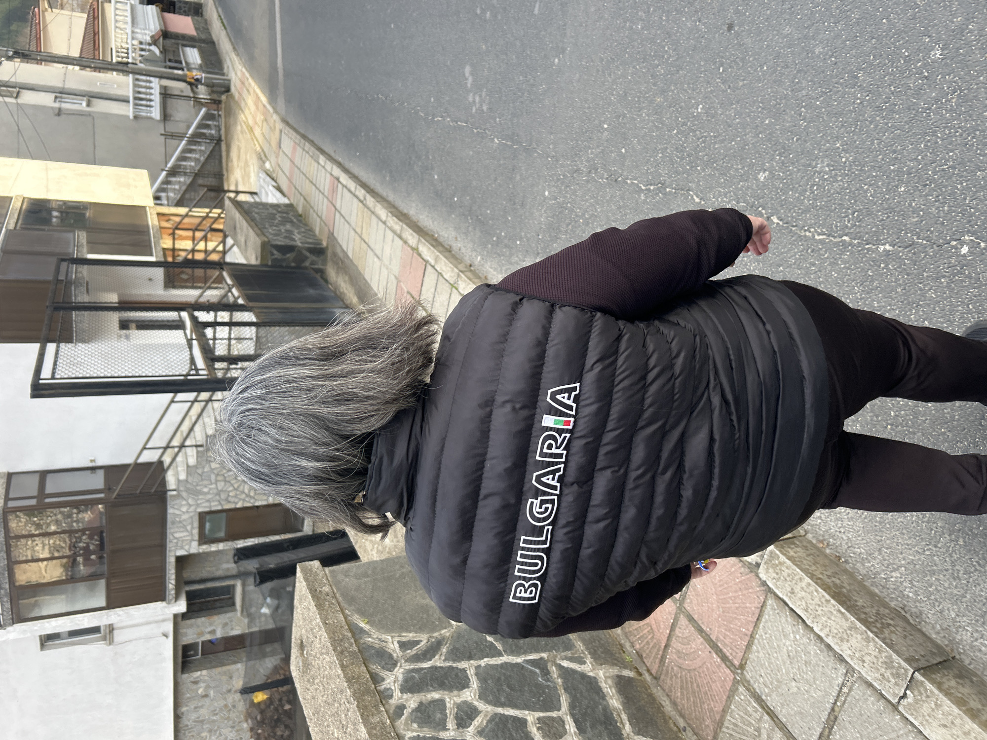 The image shows an adult with gray hair wearing a black puffy vest that reads "BULGARIA" down the back in white letters, with the "I" adorned with the Bulgarian flag. They are standing on a sidewalk in Zlatograd, Bulgaria. The sidewalk is bordered by an asphalt road on one side and a stone retaining wall on the other. Buildings are visible in the background. The person appears to be walking.