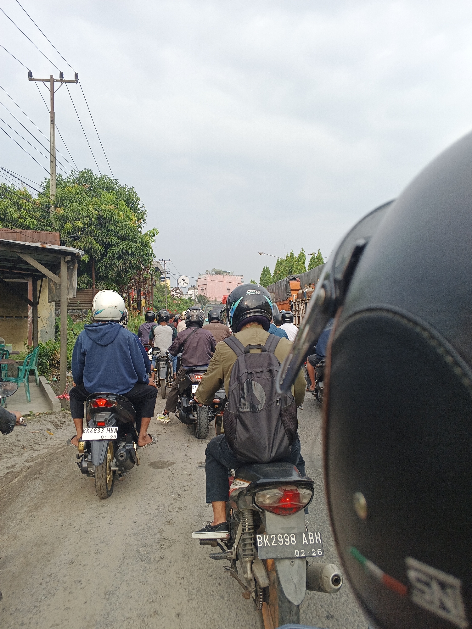 A first-person perspective shot captures a traffic jam of motorcycles on a dirt road in Tg Morawa B, Indonesia. The foreground is dominated by the back of a black motorcycle helmet, obscuring the driver's view. Ahead, a line of motorcyclists on various bikes navigate the dusty road. One rider in a blue hoodie and white helmet is prominently positioned, with a license plate reading "BK 4833 MBA". Further down the road, other riders are visible, many wearing helmets. The environment includes a utilitarian building with a tin roof and green plastic chairs on the left, and lush green trees lining the road. Overhead wires are strung from a wooden utility pole on the left. The sky is overcast.