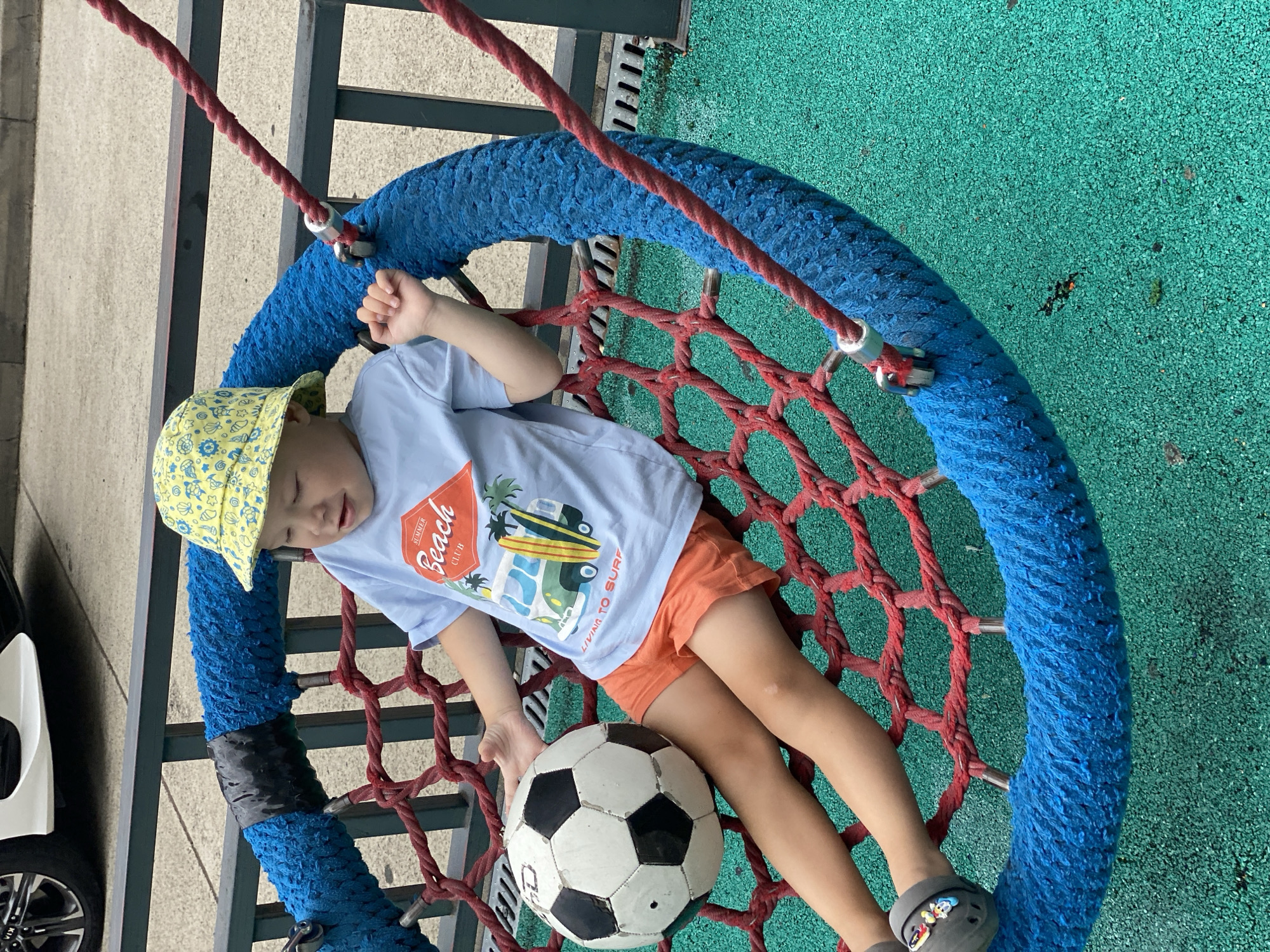 An outdoor shot shows a young boy sitting in a large, blue, netted swing. The boy is wearing a yellow bucket hat with a pattern of blue drawings, a light blue t-shirt with a "Summer Beach Club" graphic, and orange shorts. He is holding a black and white soccer ball in his left arm. The swing is suspended by red ropes and has a thick, blue, textured rim. The ground beneath the swing is covered in a green, rubberized surface, common in urban playgrounds. In the background, a dark metal fence and a portion of a white car are visible, suggesting an urban or park setting. The lighting is bright, indicating it is daytime. The scene is likely set in an urban park, possibly in Sochi, Russia, given the context.