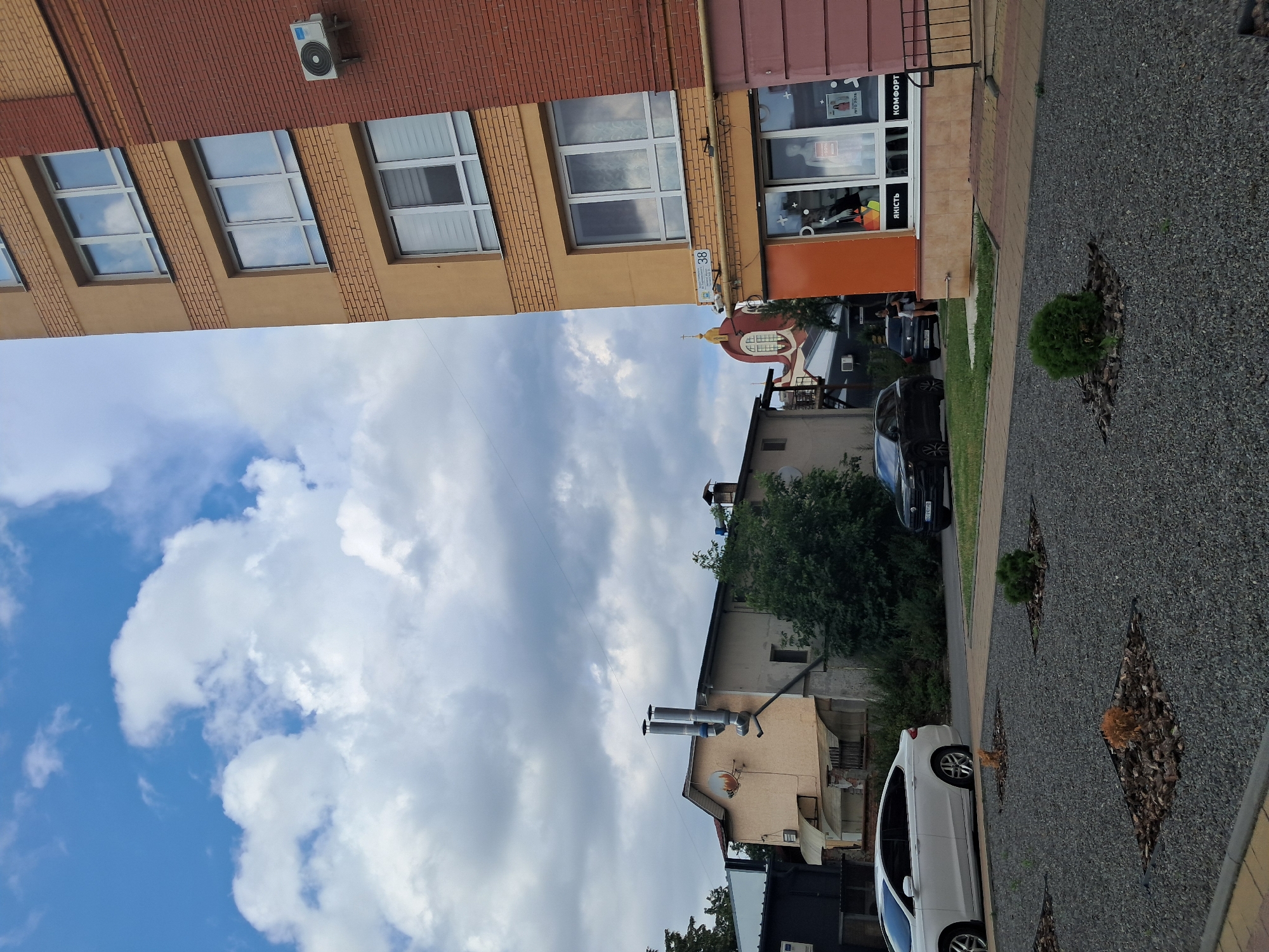 An outdoor, eye-level, medium shot captures a view of a modern apartment building with a brick facade and large windows, adjacent to a smaller, older building with two prominent exhaust pipes. The sky is partly cloudy with scattered white clouds against a blue background. In the foreground, a white sedan is parked on the left, and a dark SUV is parked on the right. The ground is covered in small gray gravel with several small, square planting beds containing decorative bark and small plants. A portion of a storefront with large windows is visible on the right side of the apartment building, displaying a mannequin. In the background, a glimpse of a building with a golden dome and a cross suggests a religious structure. The lighting indicates it is daytime.