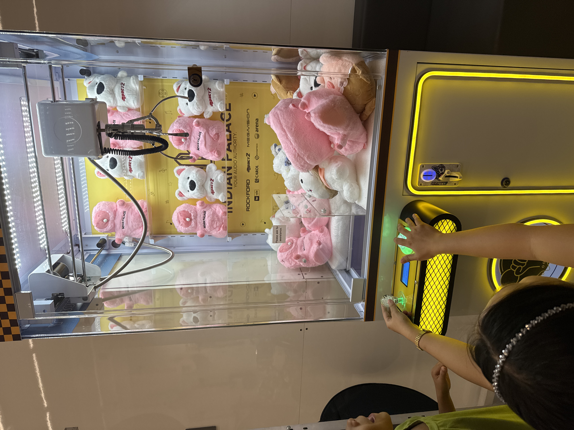 The image displays an indoor scene within what appears to be an arcade or entertainment center, focusing on a claw machine. An adult's hands are actively engaged with the machine's controls, manipulating a glowing green joystick with the right hand and holding a clear, faceted token with the left. A child, identifiable by a sequined headband and partially visible face, is looking up, seemingly observing the game.
The claw machine's transparent case is filled with an assortment of plush toys, predominantly white koala-like figures and pink pig or bunny-like characters. The interior is brightly lit, highlighting a yellow back panel adorned with text. The prominent text reads "INDIAN PALACE YOUR AUDIO AUTHORITY," with smaller brand names and logos such as "ROCKFORD," "IMPACT2," "MEGAVISION," "vybe," "CAROL," "SHOWTECH," and "arena" also visible. Several white plush toys bear the "IMPACT2" logo, while the pink ones show "INDIAN PALACE." A small white tag inside the machine contains the instruction "Please ask for assistance." The control panel area of the machine is framed by striking yellow LED strip lighting. The activity captures a moment of entertainment and focus as the players attempt to win a prize from the machine in Davao City, Philippines.