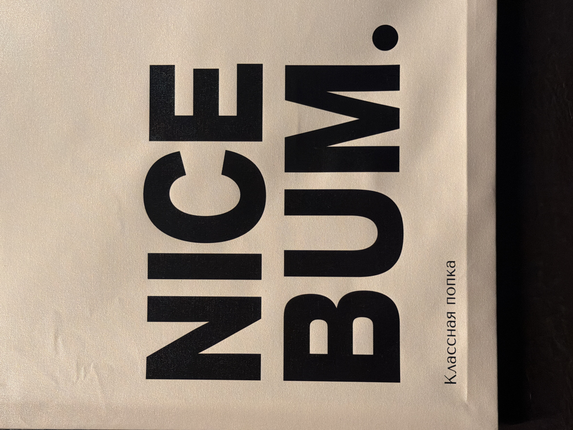 The image shows a close-up of a light-colored bag or item with the words "NICE BUM." printed in large, black block letters. To the right of the English phrase, in smaller, darker text, is the Russian phrase "Классная попка." The background is plain, suggesting the item is resting on a dark surface.