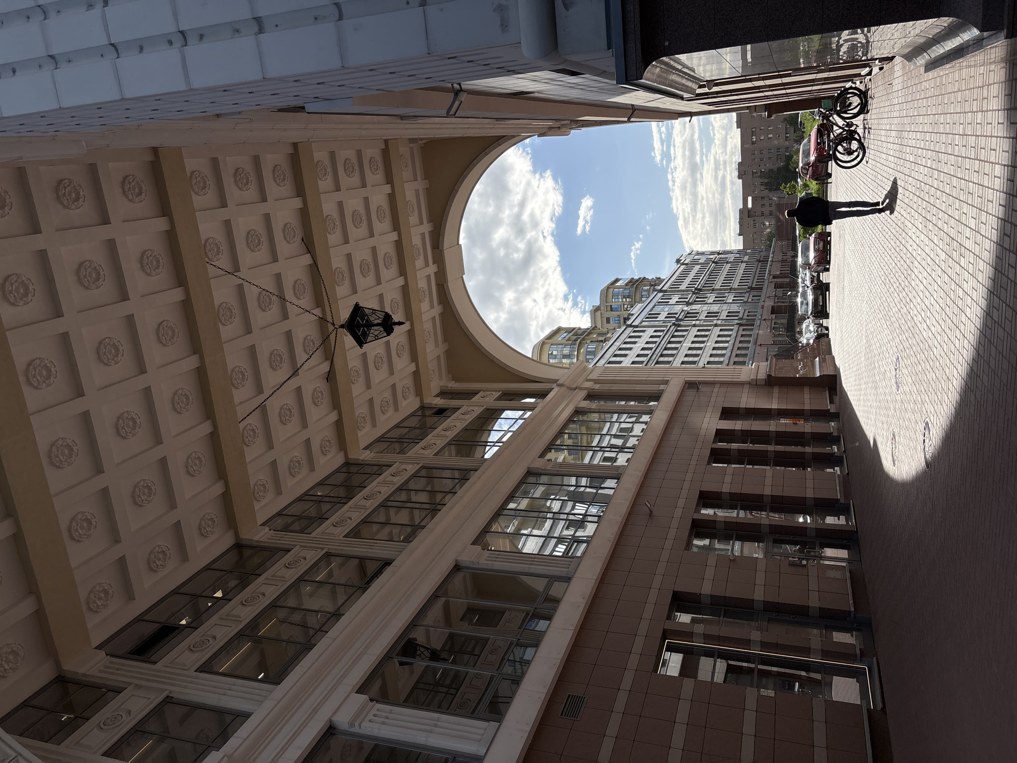 The image shows an urban view in Saint Petersburg, Russia, with a perspective looking upward through an architectural archway. The archway is ornate, featuring a coffered ceiling with decorative rosettes and a hanging lantern. The opening of the arch reveals a view of the sky with scattered clouds and sections of tall buildings with multiple windows, suggesting an area with dense urban development.
On the right side of the frame, a person is walking along a brick-paved walkway. The person is silhouetted against the sunlit bricks. Behind the person are parked vehicles and bicycles.