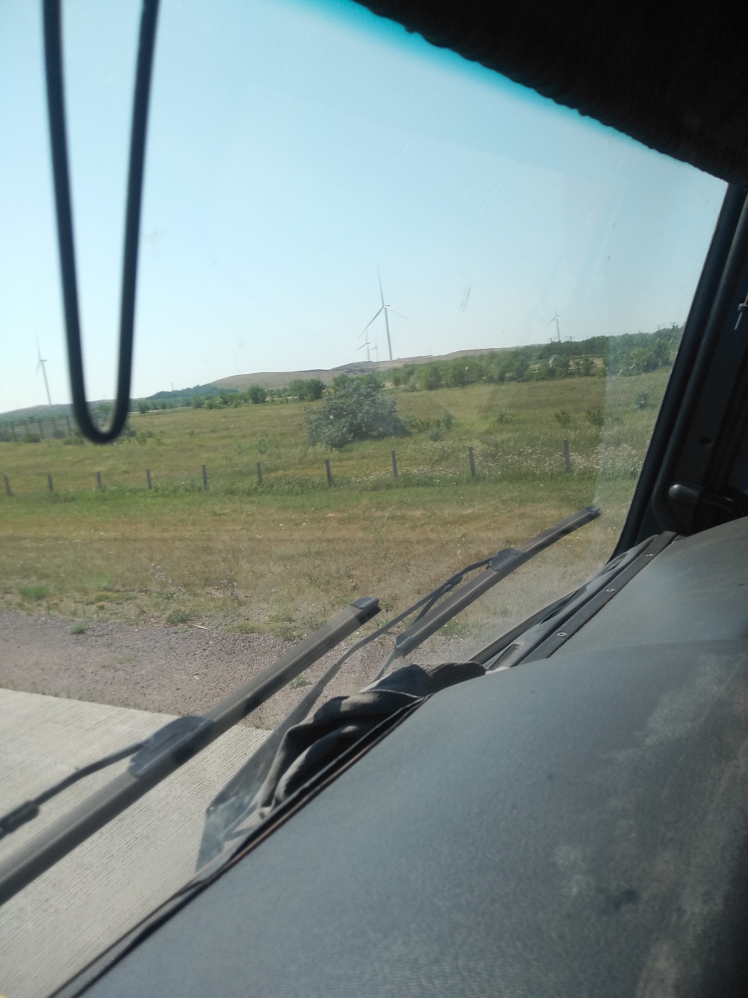 The image, taken from inside a vehicle looking out through the windshield, shows a rural landscape with wind turbines visible in the distance. The foreground consists of the vehicle's dashboard and windshield wipers. The scene is set during the daytime under a clear sky. The surrounding environment is characterized by a grassy field with scattered trees and a dirt or gravel road. The wind turbines suggest the location is likely a wind farm. There are no people or animals visible in the image. The overall impression is one of a serene, open countryside. The city of Osakarovka, Kazakhstan is mentioned, which implies this landscape is situated in or near that region.