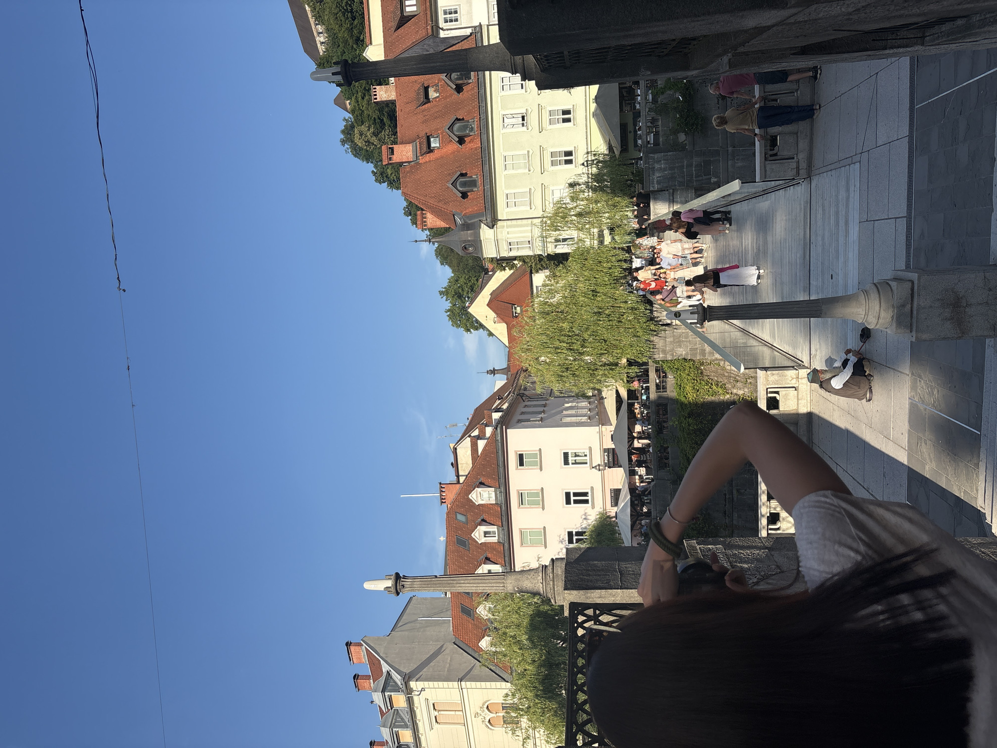 A woman with long dark hair is taking a photo with a camera in an urban setting. She is wearing a white t-shirt and has bracelets on her left wrist. In the background, a bridge spans across a waterway, with many people walking on it. Buildings with red tile roofs line the sides of the waterway. Trees with lush green foliage are visible. In the foreground, a stone pillar stands next to a dark metal railing. To the right of the bridge, two people are standing by a railing, and to the left of the bridge, a person is sitting on the ground. The sky is clear and blue.