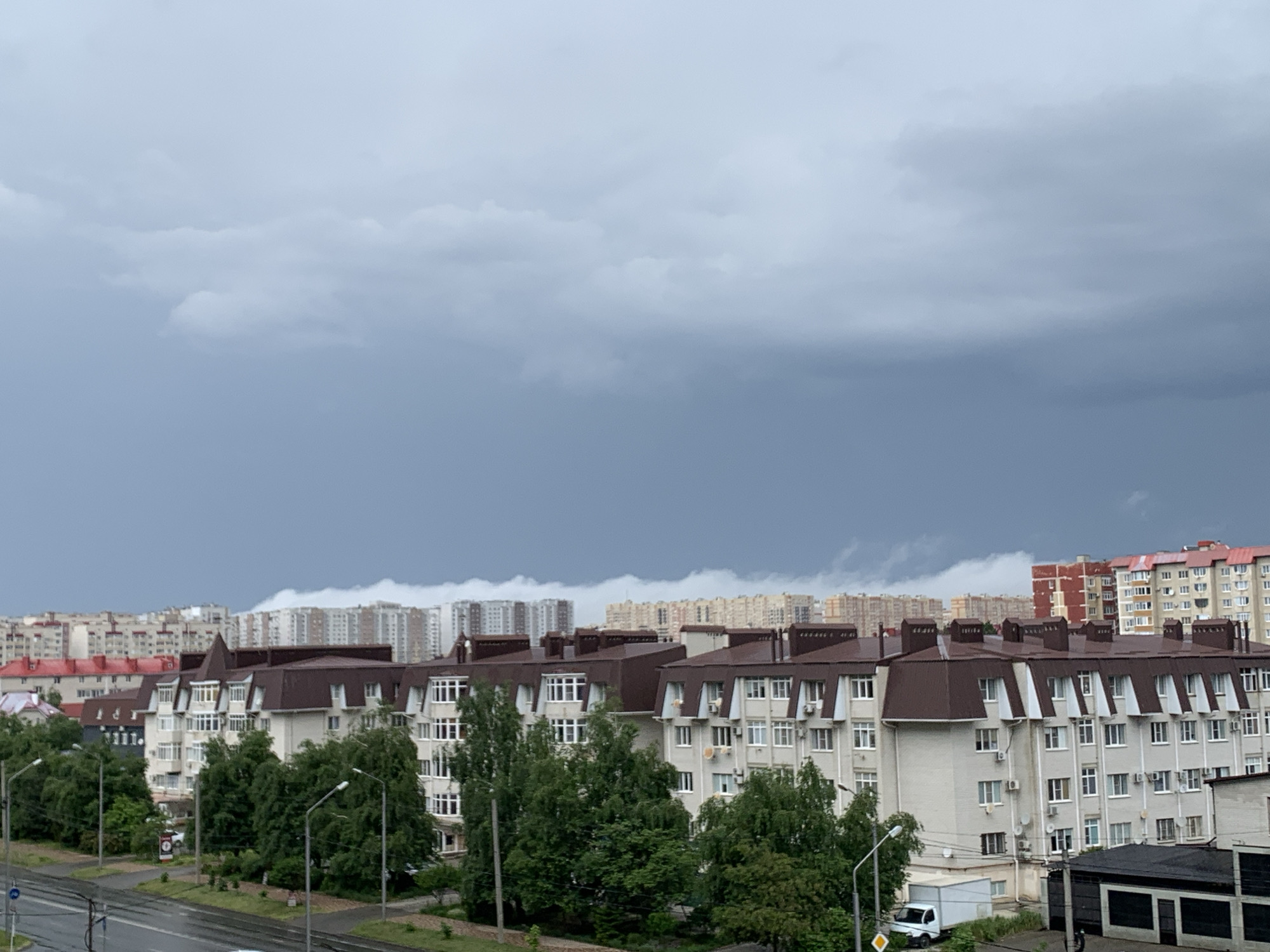 The image depicts an urban scene under a dark, overcast sky, suggesting impending rain or a recent downpour. The foreground features a wet asphalt road with white lane markings. On the right side of the road, a white van is parked. Beyond the road, a sidewalk is visible, lined with streetlights and lush green trees.
Dominating the mid-ground are several multi-story apartment buildings with brown, sloped roofs and light-colored facades. The buildings are characterized by numerous windows, air conditioning units, and balconies. Some buildings have white trim around the windows.
In the background, a dense collection of taller apartment buildings stretches across the horizon, with varying architectural styles and colors, including some with red accents. The sky is filled with heavy, dark grey clouds, creating a somber atmosphere. The overall impression is a residential area within a city, possibly on the outskirts or in a developing district, under threatening weather conditions. No people are visible in the image.