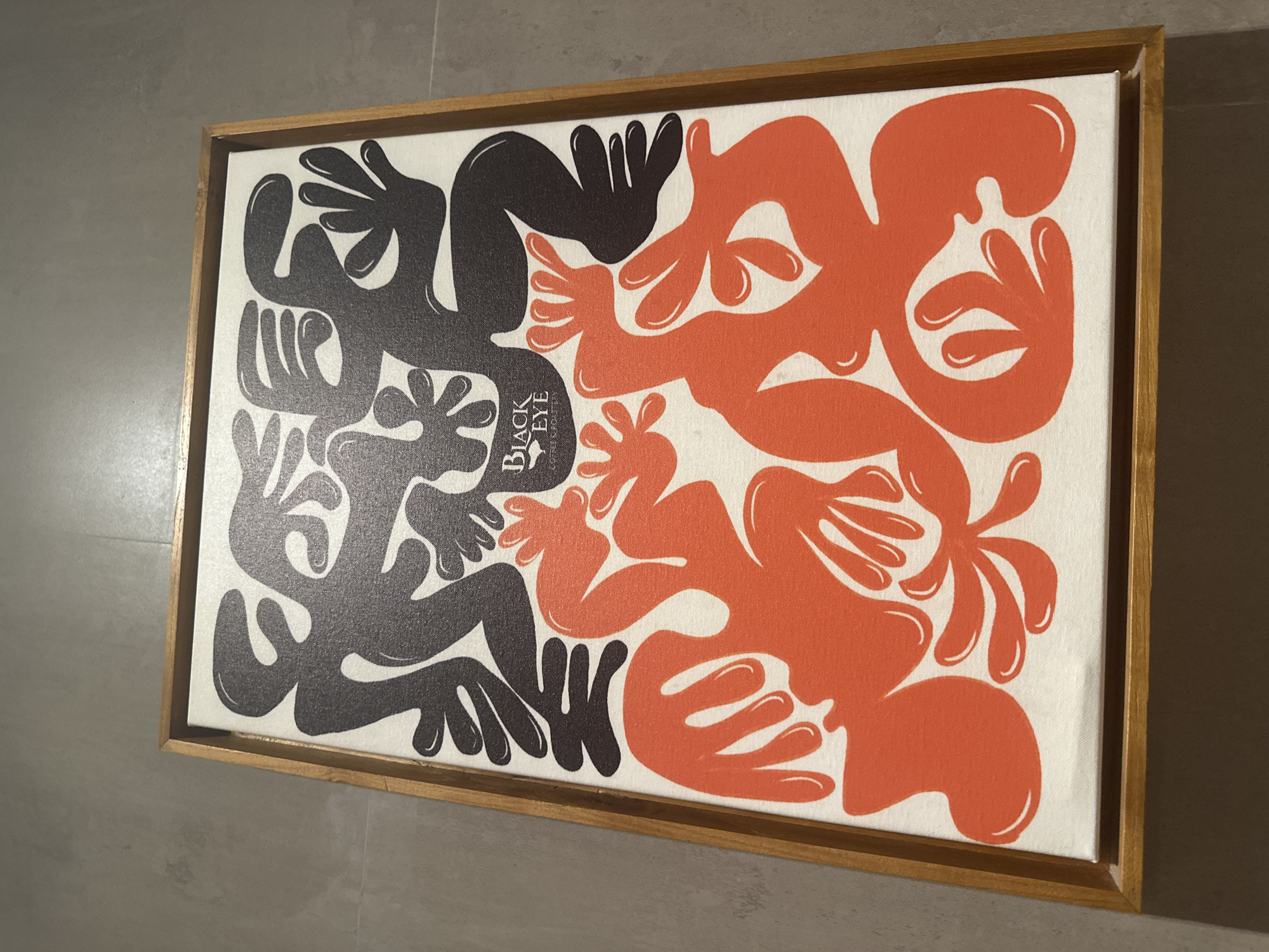 This is a close-up, overhead shot of a framed abstract artwork on a white canvas. The artwork features two distinct layers of stylized figures. The top layer is rendered in solid black, with undulating, organic shapes that suggest limbs and bodies in motion. The bottom layer is in a vibrant orange, with similar abstract forms that interlock and complement the black figures. In the upper center of the canvas, a small logo with the text "BLACK EYE" above smaller text that reads "COFFEE & ROASTERY" is visible in white. The canvas is set within a thin, light-colored wooden frame. The artwork is positioned on a grey, textured floor, which appears to be concrete. The overall lighting is neutral. The image does not depict any people or specific location in Ubud District, Indonesia.