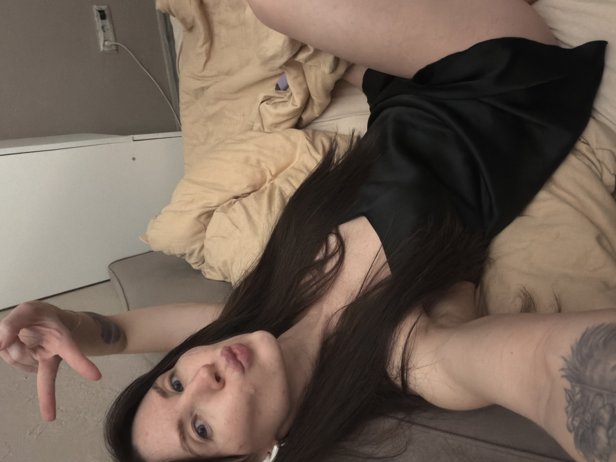 A woman with long dark hair lies on a bed covered with a light brown blanket. She wears a short black dress. Her left arm is extended, with her fingers forming a peace sign. Tattoos are visible on both arms. The bed is situated in a room with white furniture and a wall-mounted electrical outlet. There is no evidence to suggest the location is Perm, Russia.