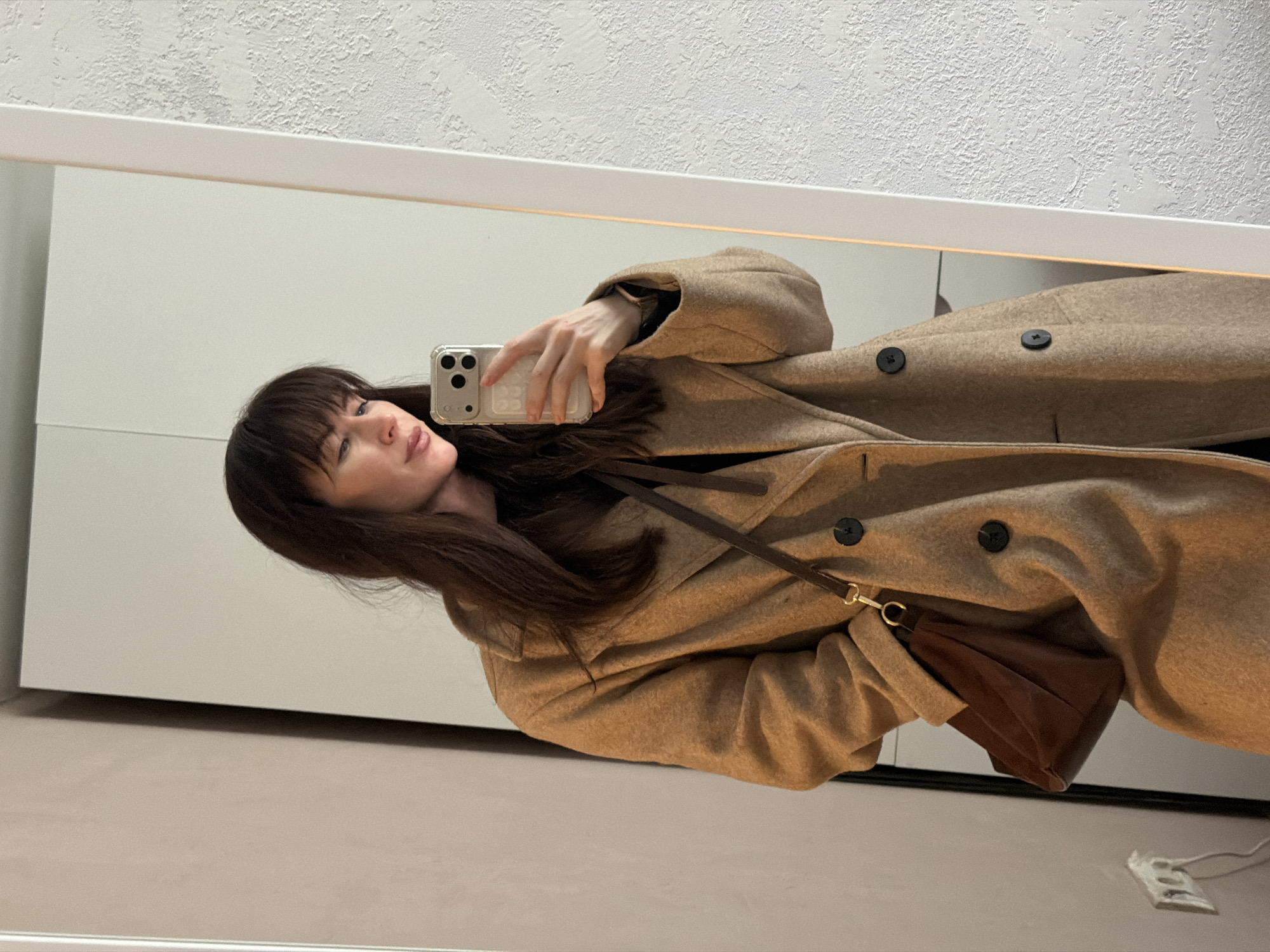 A woman with dark hair and bangs is taking a selfie in a mirror. She is wearing a tan, double-breasted coat with large buttons and a brown leather purse with a strap. The background includes a white wall and a textured white ceiling. The photo appears to be taken indoors, possibly in a home or apartment. There is no indication of Perm, Russia in the image.