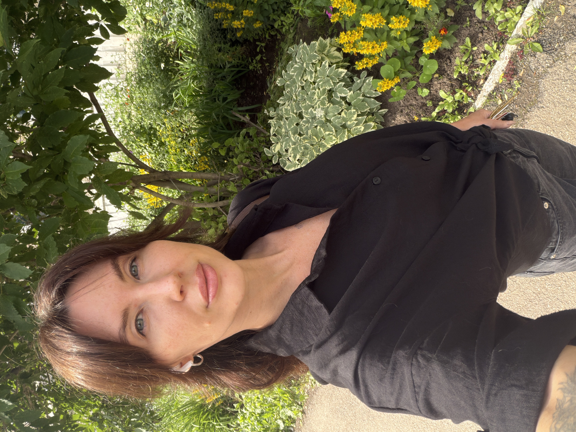 A woman with brown hair and blue eyes is taking a selfie outdoors in a park or garden setting. She is wearing a black button-up shirt and dark jeans, with white wireless earbuds in her left ear. In the background, there are various green plants, bushes, and yellow flowers. The ground appears to be a path made of small stones or gravel, with a concrete border. A tree with green leaves is visible to the left of the woman, casting shadows. The lighting suggests it is daytime with sunlight. The specific location in the city of Perm, Russia, is not identifiable from the image alone.