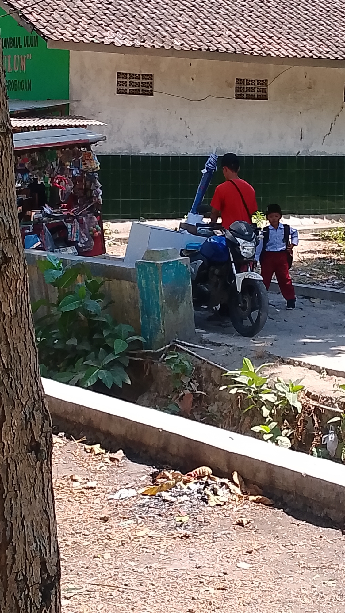 An overhead shot captures a street scene in Karanglangu, Indonesia. A person in a red shirt stands by a blue motorcycle, with a child in a school uniform nearby. The background features a building with a tiled roof and green tiled walls. To the left, a stall displays various items. The ground is covered with dirt and scattered leaves.