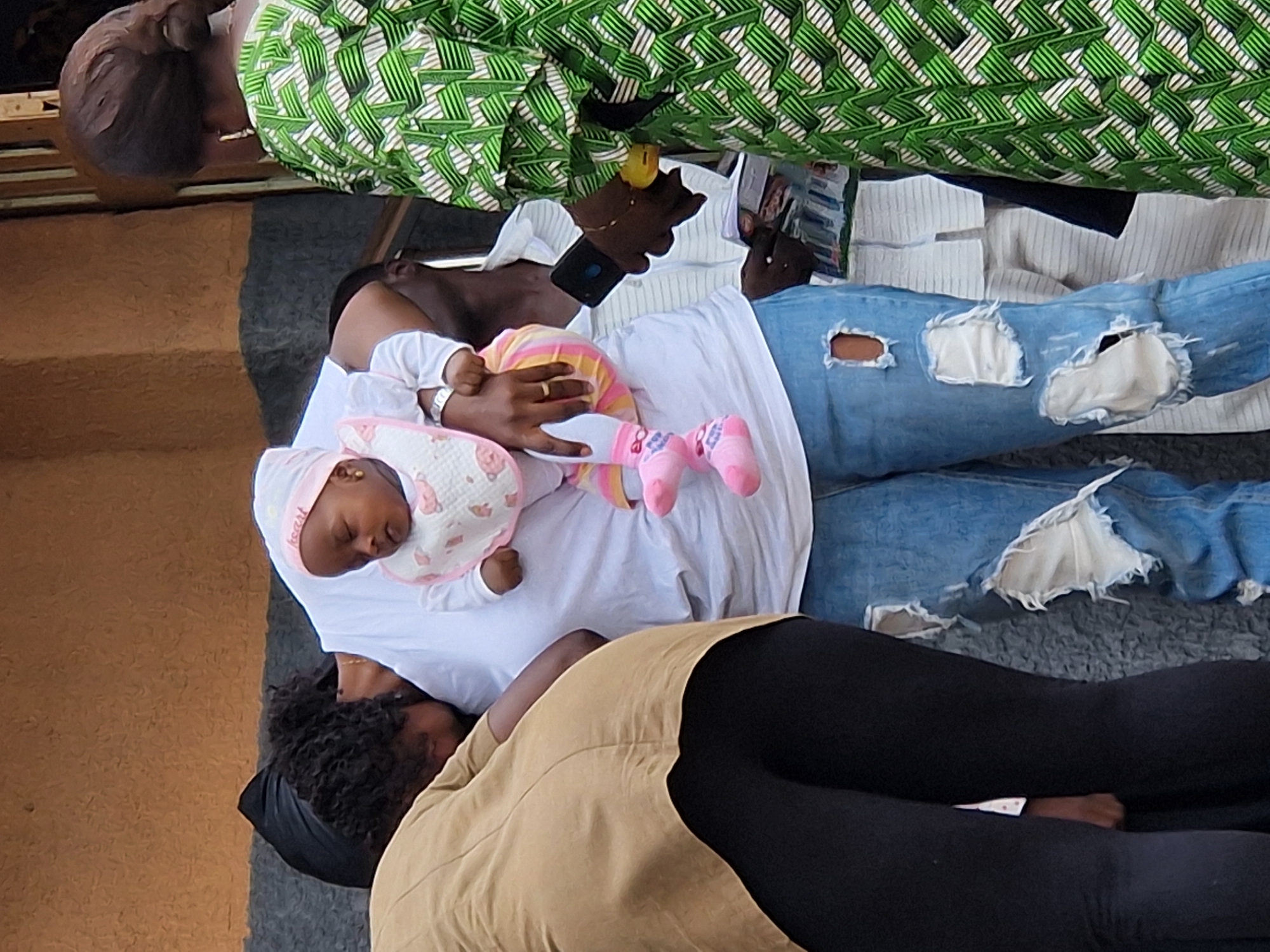 A close-up, medium shot shows several people gathered together outdoors, with a focus on a baby being held. The baby, wearing a pink and white outfit and a bib, is cradled in the arms of a person who is mostly out of frame, but whose ripped blue jeans are visible. To the left, a person in a tan jacket and black pants is mostly seen from the back. To the right, a person in a green and white patterned dress stands looking away from the camera. Another person in a white shirt is partially visible behind the person holding the baby. The background appears to be a building with textured walls in shades of brown and gray. The overall lighting suggests it is daytime.