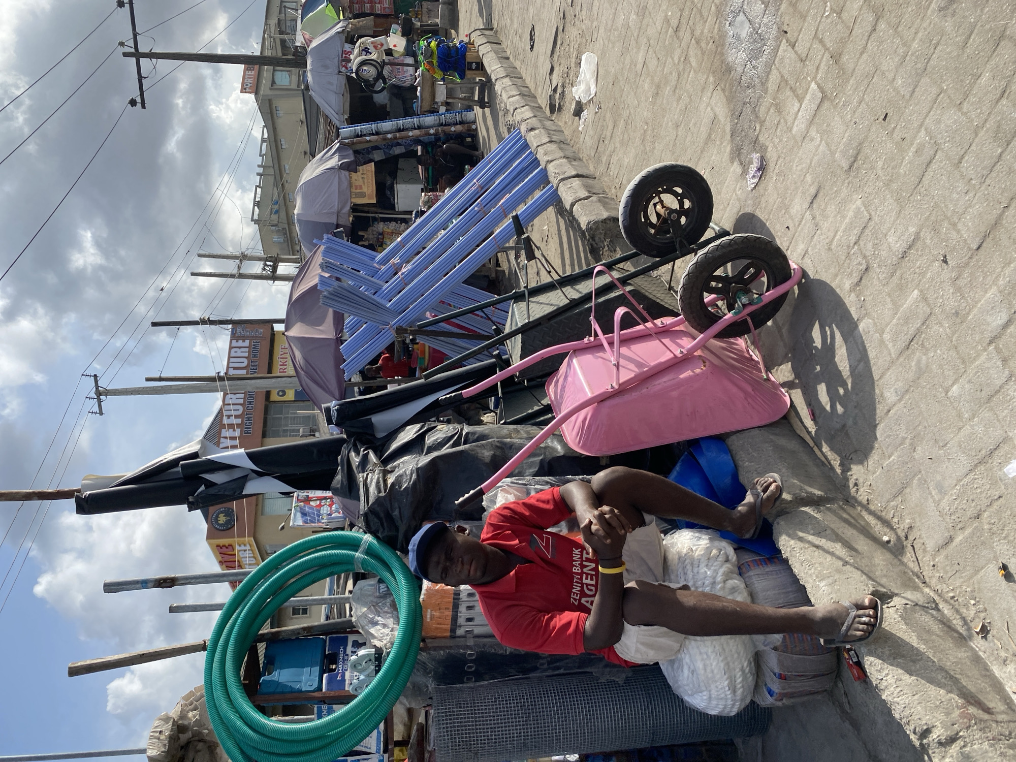 A man sits next to a sidewalk on what appears to be a pile of wares in Lekki, Nigeria. He is wearing a red t-shirt, blue cap, and sandals. The shirt has "ZENITI BANK AGENT" printed on it. The wares next to him include a coiled green hose, mesh fencing, and bags of materials. Behind him, there's a pink wheelbarrow and blue pipes on a cart. In the background, there is a store with signage including "FURE YE FURTURE RIGHT CHOIC VEET HOME RKİYE." There are also telephone poles and overhead wires, as well as umbrellas covering various market stalls. The street is paved with brick-like pavers, and the sky is overcast.