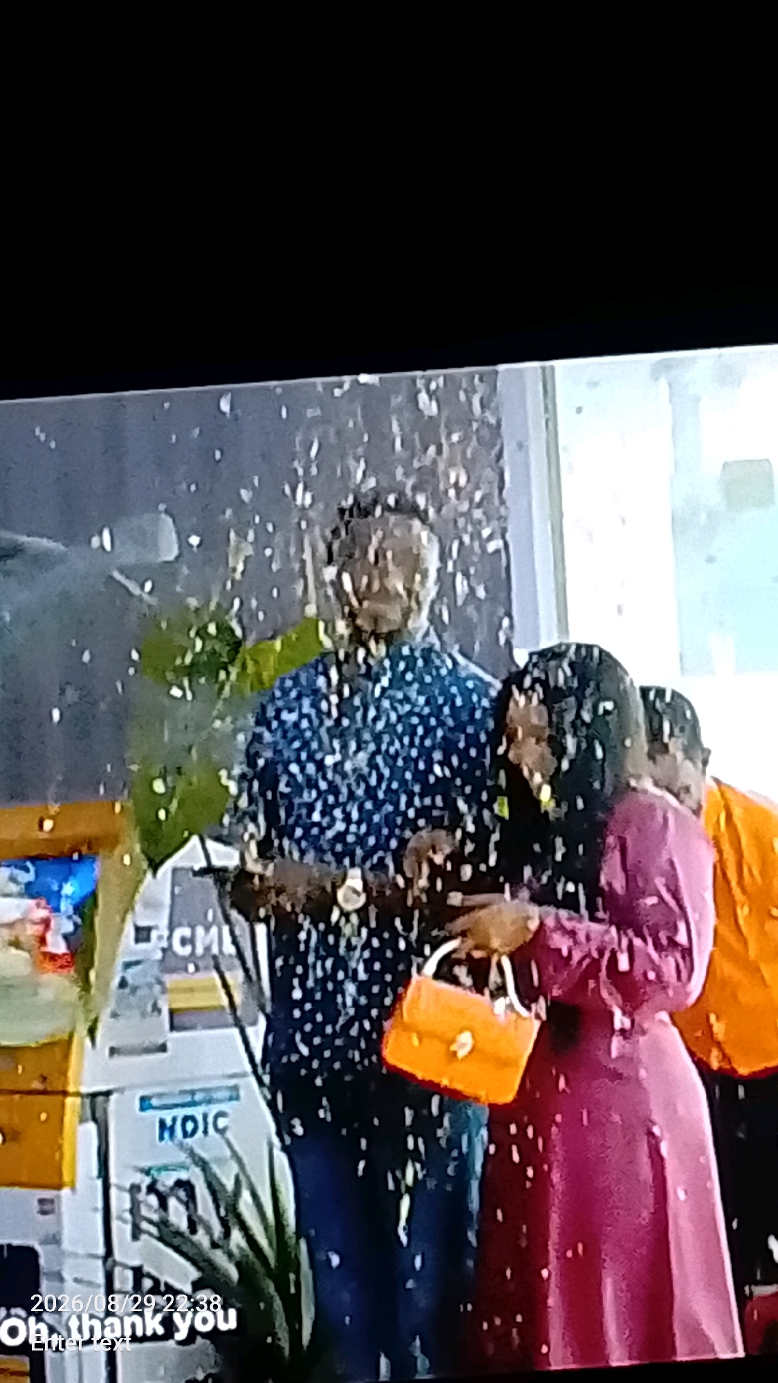 In Lokoja, Nigeria, a man wearing a blue patterned shirt stands with his arm around a woman dressed in a magenta dress. The woman holds a small orange handbag. A third person, partially visible in an orange garment, stands to the right. Confetti or glitter is falling around them, suggesting a celebratory occasion. In the background, there's a green leafy plant and signs with text, including "HDIC" and "CM." The scene appears to be indoors, possibly at an event or gathering.