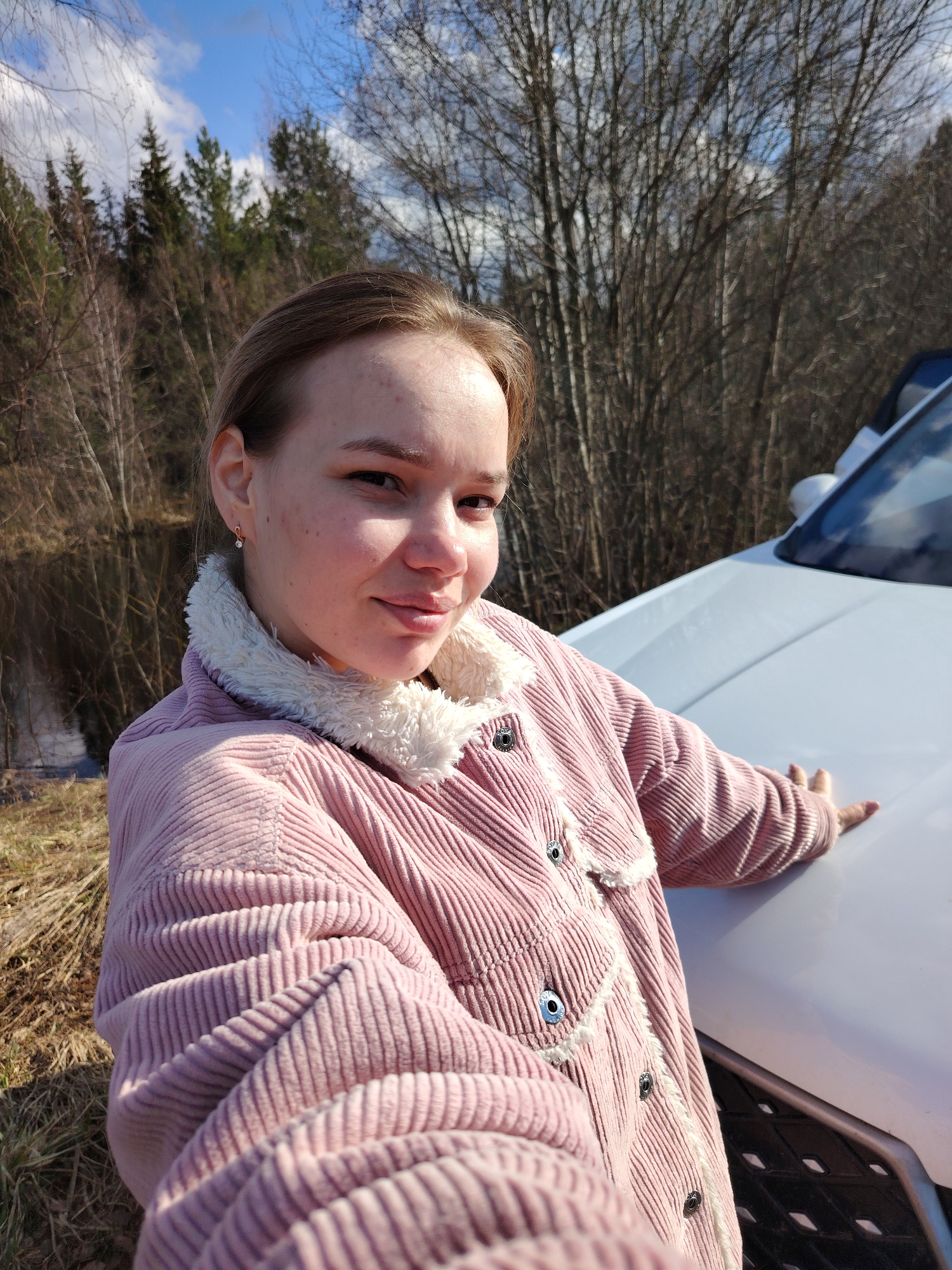 The image is a medium shot selfie of a young woman outdoors in Chaikovsky, Russia. She is leaning on the hood of a white car. The woman has fair skin, light brown hair pulled back, and is wearing a pink corduroy jacket with a white faux-fur collar and trim. A small earring is visible in her left ear. Behind the woman and the car, there is a body of water, possibly a lake or river, with a wooded area in the background. The trees are mostly bare, indicating either late fall or early spring. The sky is partly cloudy.
