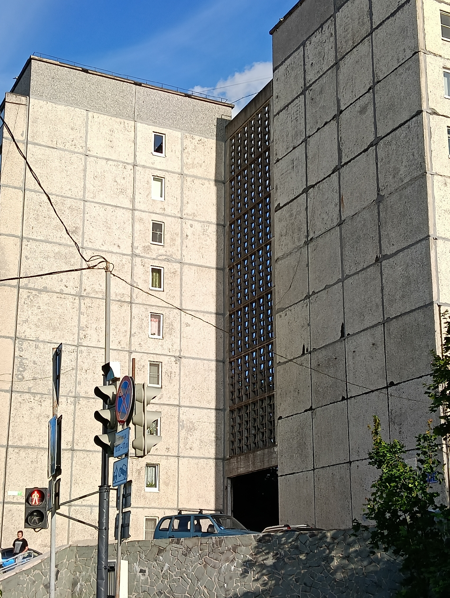 The image depicts a street view in Petrozavodsk, Russia, focusing on a concrete building with a distinctive grid-like window structure. A traffic light is visible in the foreground, displaying a red pedestrian signal, with a person standing near it. The building itself appears to be an apartment block, characterized by its modular concrete panels and numerous small windows. To the right, a portion of a car is visible entering a covered area or garage beneath the building. The scene is set against a backdrop of a partly cloudy blue sky.