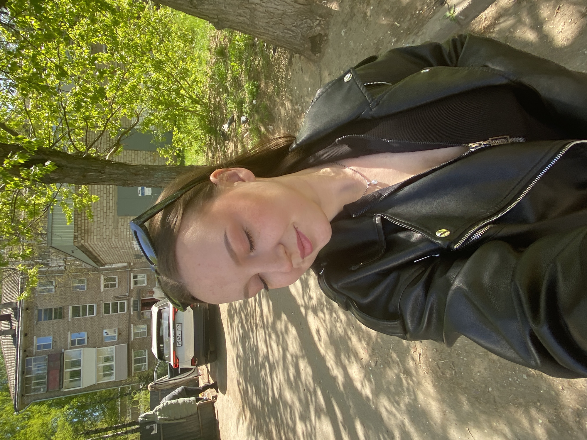 A person is standing outdoors on a paved surface, potentially a sidewalk or street, in a residential area of Chaikovsky, Russia. The person is wearing a black leather jacket and a necklace. A multi-story brick apartment building is in the background, with a white SUV parked nearby. Trees and other vegetation are present, creating dappled shadows on the ground.