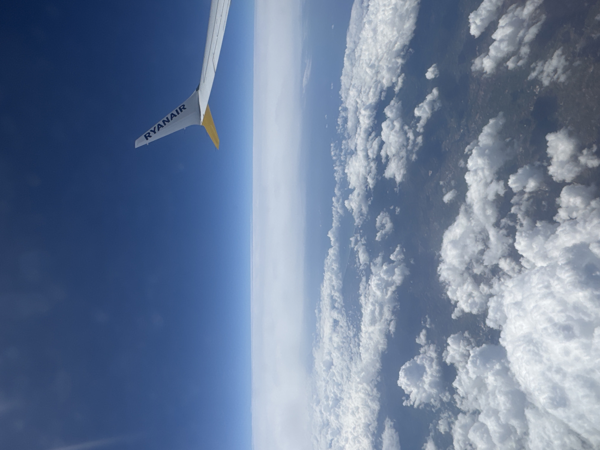 The image presents a view from an airplane window. In the upper left corner is a portion of a Ryanair airplane wing, identifiable by the company's name printed on it, and the yellow winglet at the tip. Adjacent to the wing, there is a clear, deep blue sky. The horizon is slightly hazy, with a white or light gray band running along it. Below the horizon is a view of the earth, partially obscured by a dense layer of white, fluffy clouds. Below the clouds, hints of land are visible. The specific location is unidentifiable.