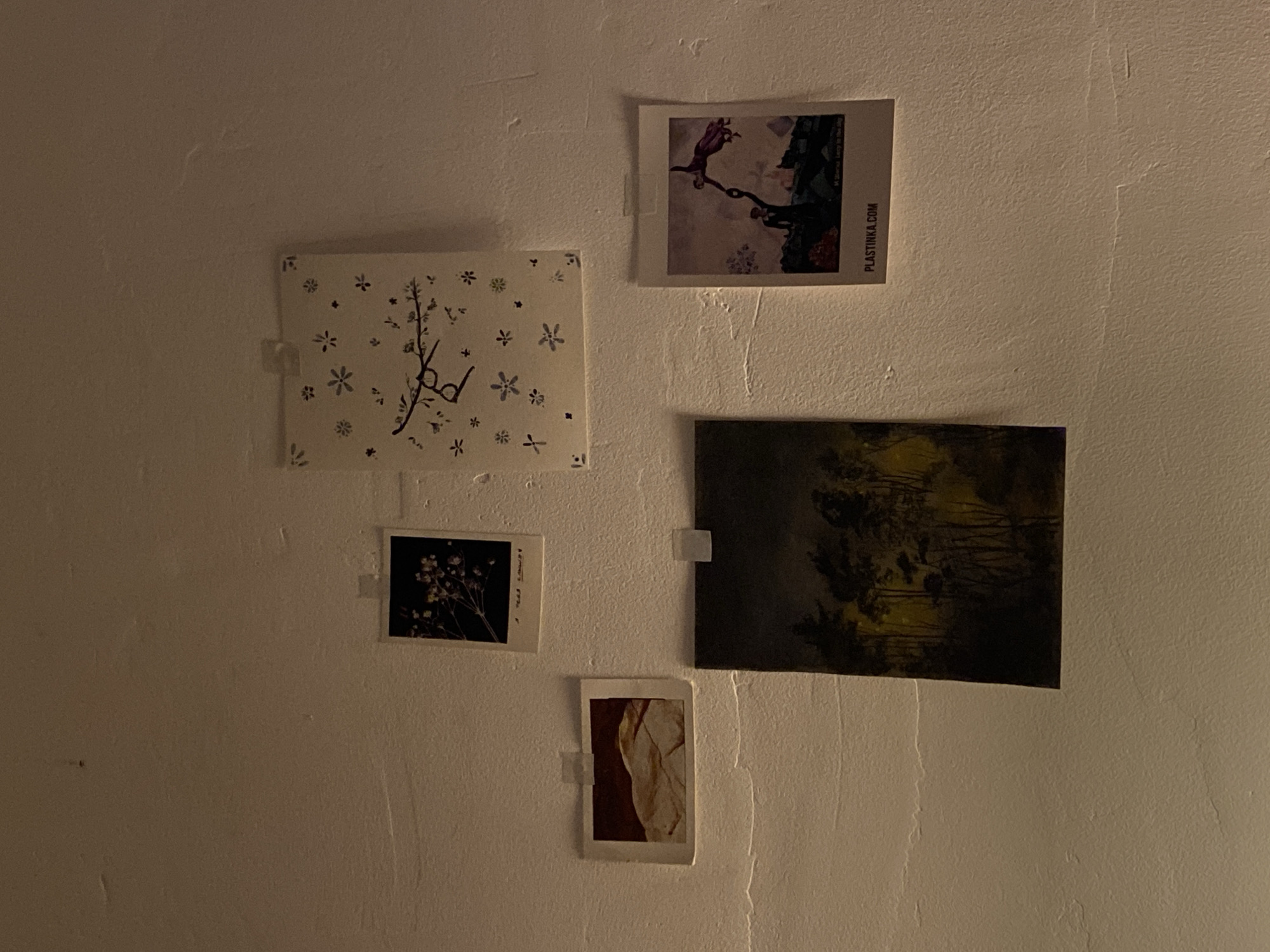 Five small pictures are taped to a white wall. From top left to bottom: A square white picture with black floral designs and a pair of glasses. Next is a rectangular picture with people in a fantasy scene. Below the floral picture is a small square picture with a floral design. To the right is a rectangular landscape picture. Below the landscape picture is a small vertical rectangular picture with a cream and reddish-brown abstract design. The location of the image is not apparent.