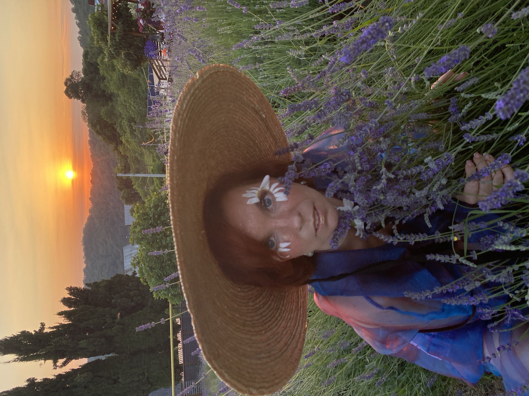 A woman with reddish-brown hair, wearing a wide-brimmed straw hat and a flowing blue and red garment, is seated in a field of lavender during sunset. She has daisy petals affixed to her face, one over her right eye and two on her cheeks. Her hands are visible among the lavender stalks. In the background, hills are covered with trees, and a warm, orange sky indicates the setting sun.
