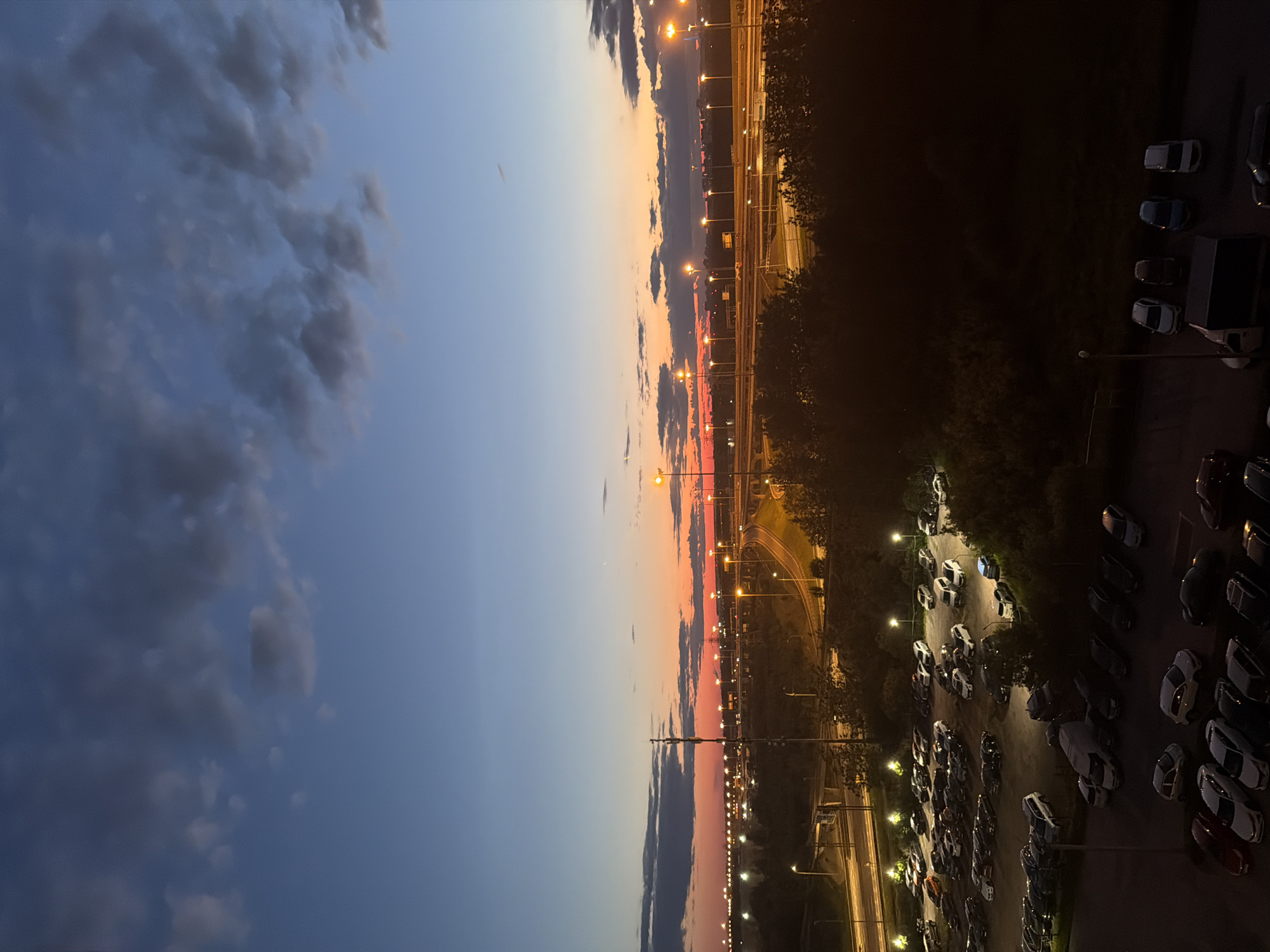 The image depicts a twilight scene over a parking lot and a highway. The sky displays a gradient of colors, transitioning from deep blue at the top to vibrant orange and red near the horizon, indicative of sunset or sunrise. Scattered clouds are visible in the upper portion of the sky.
In the mid-ground, a multi-lane highway is illuminated by streetlights, with the glow of distant city lights also visible along the horizon. A tall communication tower stands prominently on the left side of the highway. A large parking lot is situated in the foreground, filled with numerous parked cars. The vehicles are varied in size and color, and their headlights or interior lights are not illuminated, suggesting they are parked and the area is lit by overhead lamps. Trees and bushes line the edge of the parking lot and the highway, casting dark shadows.
The lower portion of the image shows a closer view of some parked cars, including sedans, SUVs, and a van. The lighting is dim, with artificial light sources creating localized bright spots around the parked vehicles and along a nearby road. The overall impression is an urban or suburban landscape during the transitional hours of twilight.