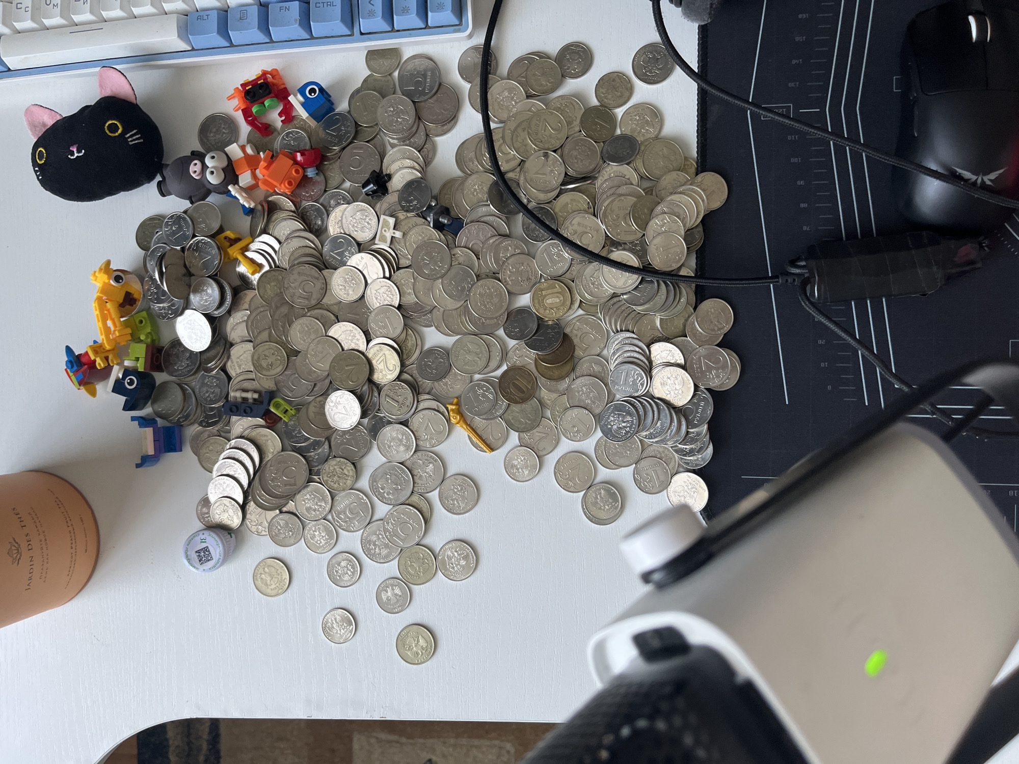 The image depicts a white table surface with various objects scattered across it. In the upper left corner is a cylindrical container of tea. To the right of the tea container, there is a collection of Russian coins, predominantly silver in color, piled and spread out. Among the coins, several small colorful toy figures, resembling Lego-style building blocks, are placed. A plush black cat toy with pink ears is also visible near the top right of the image, next to a portion of a white computer keyboard with blue keys. In the lower portion of the image, a white electronic device with a green light is positioned, and a black computer mouse is visible on a black mousepad with grid markings. The scene does not contain any information to suggest a specific location within the city of Saratov, Russia.