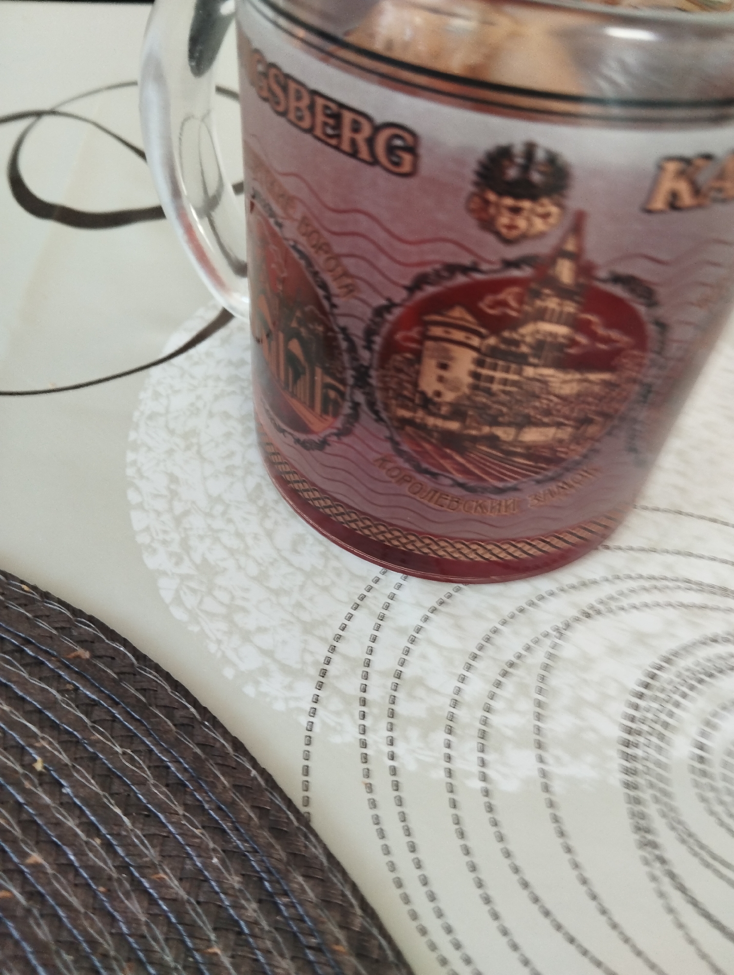 A close-up shot shows a mug with the text "KÖNIGSBERG" printed on it, possibly referencing the former German city now known as Kaliningrad. The mug features a design of a castle or fortress, with text below reading "КОРОЛЕВСКИЙ ЗАМОК" which translates to "Royal Castle." The mug sits on a patterned surface, and a part of a wicker placemat is visible in the lower-left corner. The environment suggests an indoor setting, likely a table or countertop.