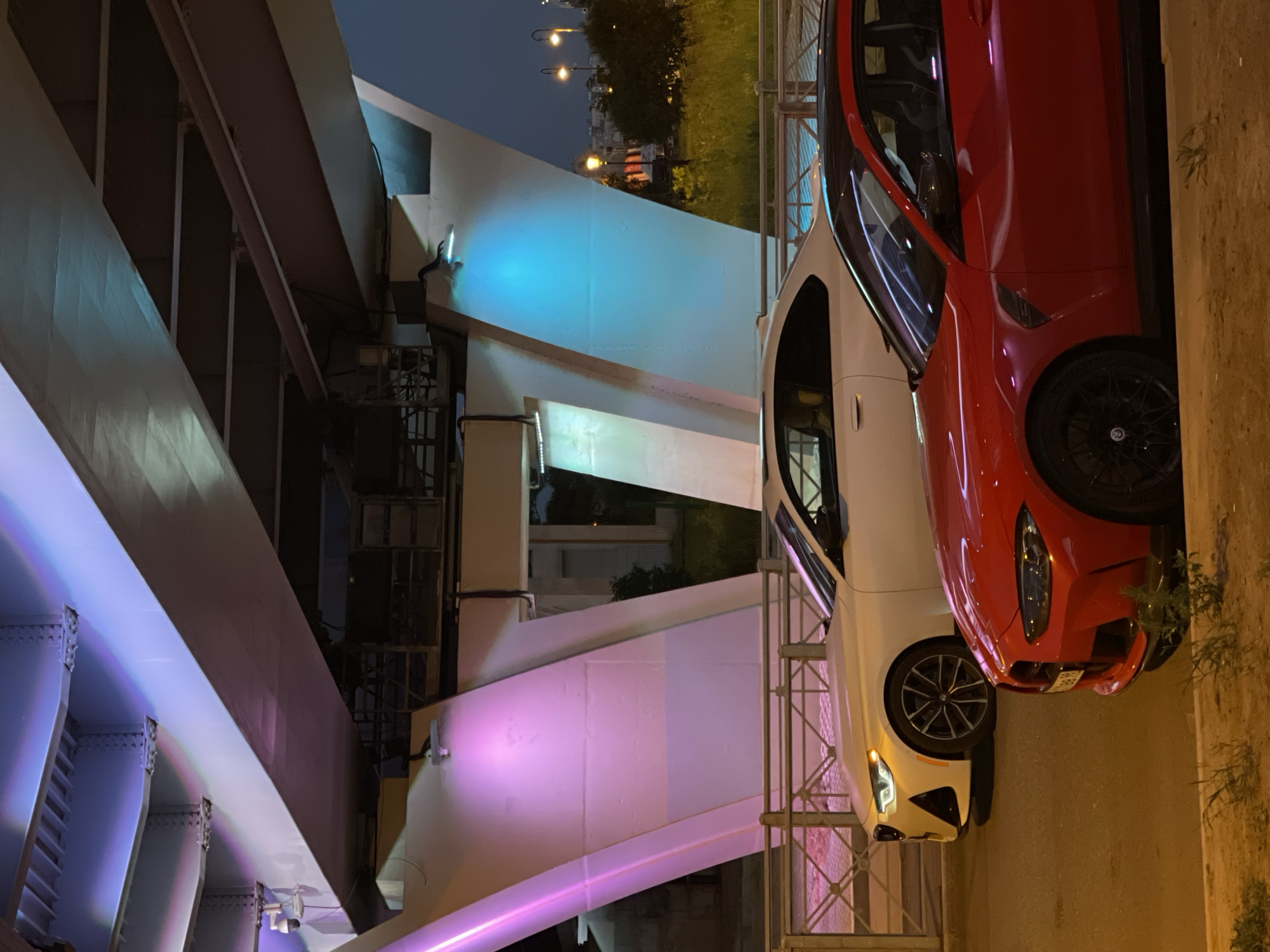 The image captures two BMW cars parked side-by-side on a street at night, beneath a large illuminated bridge. A white BMW coupe is parked in the background, and a red BMW coupe is in the foreground. The bridge is lit with vibrant purple and blue lights, casting colorful reflections on the cars and the surrounding concrete structures. Streetlights and distant buildings are visible in the background, suggesting an urban setting. The lighting creates a dramatic and modern atmosphere.