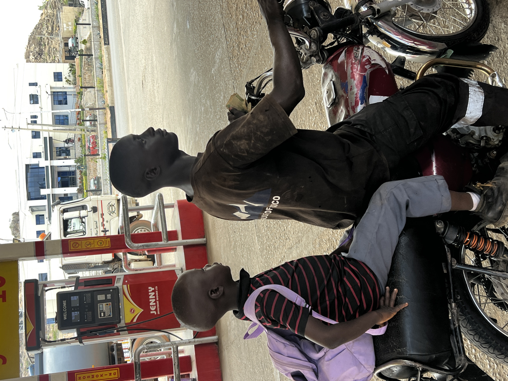 This image captures a scene at a petrol station in Jos, Nigeria, showing two males, likely an adult and a child, on a red motorcycle. The image is oriented vertically, showing subjects from a side profile looking upwards.
The man, appearing to be an adult or older teenager, sits on the motorcycle, wearing a dark, possibly stained, t-shirt with "ULFOF NICO" partially visible print, and dark trousers with reflective strips on the lower legs. He is holding money in his left hand, suggesting a transaction for fuel. Behind him, a young boy sits on the motorcycle's passenger seat, wearing a red and black striped t-shirt and light grey trousers, with a light purple backpack on his lap. Both individuals are looking intently upwards and to the left of the frame.
The motorcycle is red with silver accents, appearing well-used, with some paint wear visible on the tank. To the left, a red fuel pump is prominently featured, displaying "WELCOME" and numerical readings "465000" and "358". Text on the pump clearly identifies it as belonging to "JENNY INVESTMENT NIGERIA LTD". A "NOSMOKING" sign is also visible. Behind the fuel pump, a white truck with "TRUCK New Special" written on its side is parked.
In the background, there are several multi-story buildings, mostly white with blue-tinted windows, indicating an urban or semi-urban setting. Further in the distance, a large rocky hill is visible under what appears to be bright, clear daytime conditions. The overall scene suggests an ordinary day at a bustling petrol station.