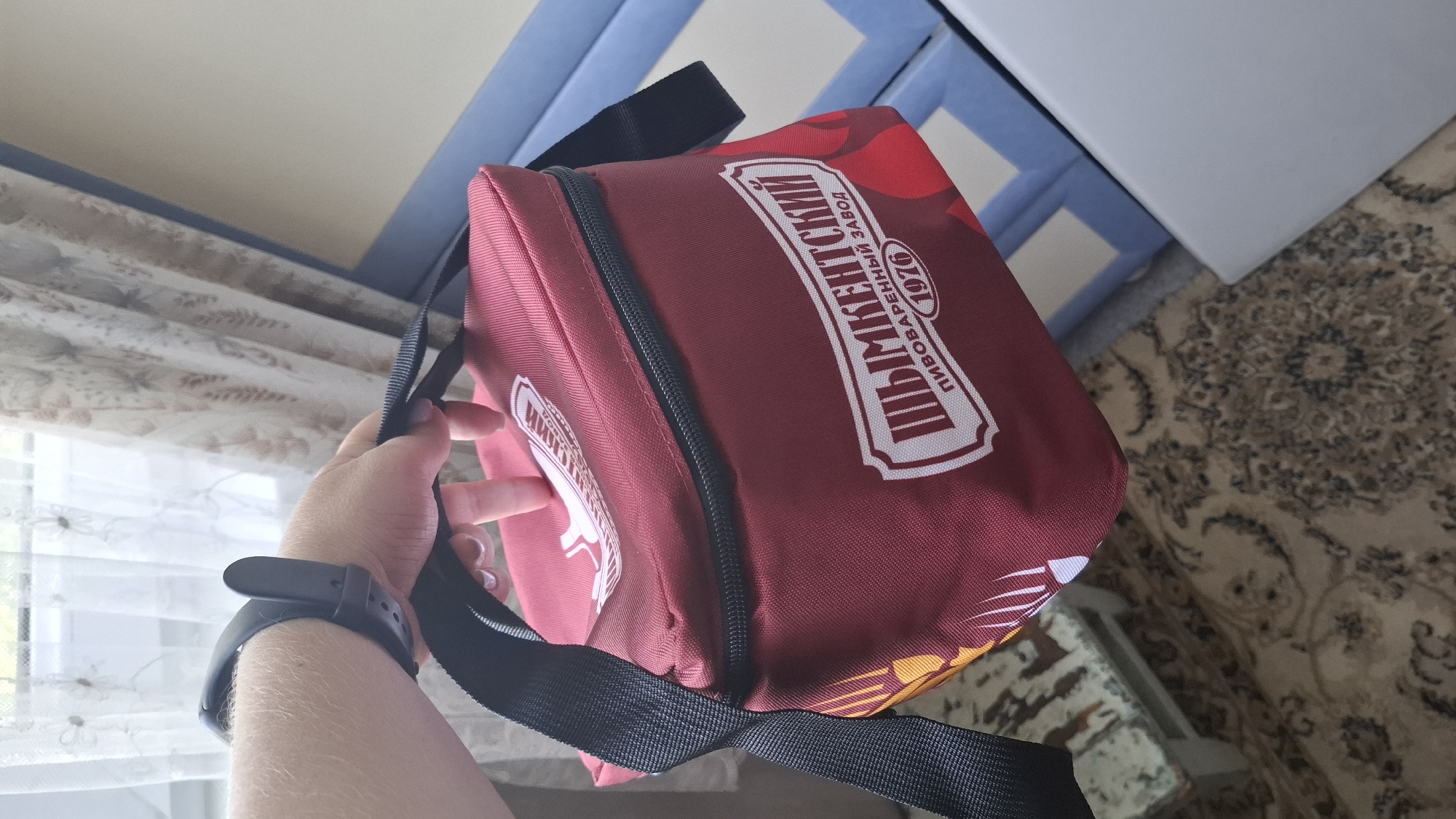 A person's hand wearing a black fitness tracker is holding a maroon insulated bag with a black strap. The bag has white lettering that reads "ШЫМКЕНТСКИЙ ПИВОВАРЕННЫЙ ЗАВОД 1976", which translates to "Shymkent Brewery 1976". The background shows a window with sheer curtains and a patterned carpet.
