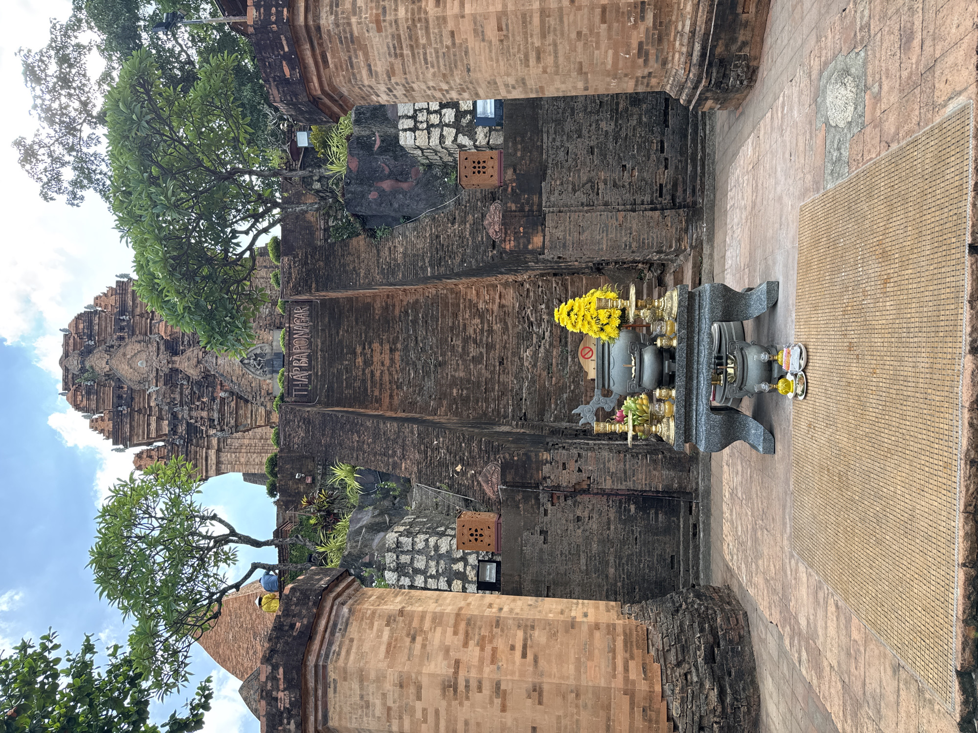 This image captures a view within a historic brick structure, likely a temple complex, in Bac Nha Trang Ward, Vietnam. The scene is dominated by weathered brick walls and large, ancient-looking brick columns. In the foreground, a dark stone altar holds offerings, including a prominent bunch of bright yellow flowers, a small statue, and other decorative items. To the right of the altar is a tan-colored mat with a grid pattern, possibly for seating or display.
In the background, a towering brick structure with intricate architectural details rises against a bright blue sky with scattered clouds, suggesting it is daytime. Lush green trees with full foliage are visible, one arching over the scene from the left, and another to the right, casting shadows and adding a natural element to the ancient setting. In the distance, on the left side of the frame, two people can be seen, one wearing a blue shirt and the other a yellow shirt, offering a sense of scale and suggesting visitors exploring the site.
A sign is partially visible on a brick wall to the right of the frame, with text that appears to read "THAP BARONAGAR". This is a strong indicator of the location, as Thap Ba Ponagar is a well-known Cham temple complex in Nha Trang. The overall impression is one of historical significance, spiritual atmosphere, and a blend of ancient architecture with natural beauty. The lighting and clear sky suggest fair weather.