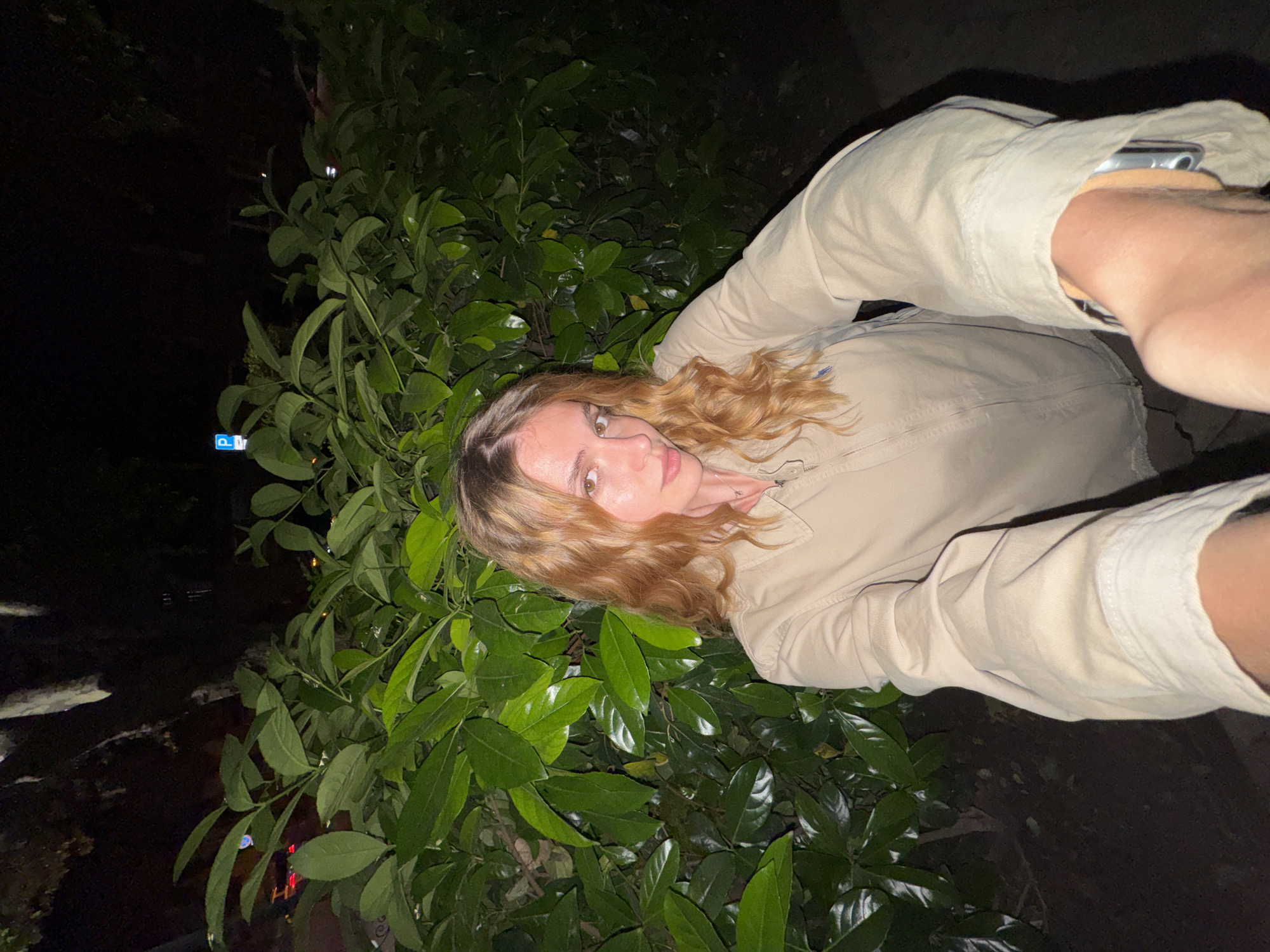 A woman is taking a selfie in front of a bush with large green leaves. She is wearing a light-colored shirt with long sleeves and is holding a phone in her right hand. The image is taken at night, and the background appears to be dark. A parking sign is visible in the background. The exact location in Tbilisi, Georgia cannot be determined from the image.