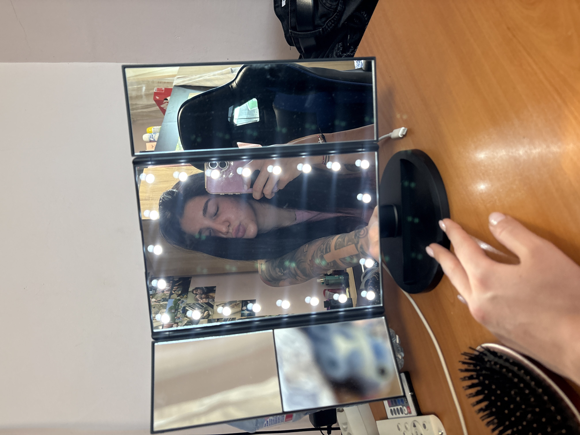A photograph shows a lighted, folding vanity mirror reflecting a person taking a selfie. The mirror is sitting on a wooden surface and a person's hand is visible near the mirror base. A hairbrush is positioned beside the hand. Reflected in the top mirror segment is a black gaming chair and a portion of a black bag. The selfie depicts a person with dark hair, an arm tattoo, and a pink top. Smaller mirrored squares below this reflect a wall of pictures and parts of the room. The presence of Cyrillic script on the power strip near the mirror implies the photograph may have been taken in a country such as Bulgaria; it is not possible to confirm from this picture that it was taken in the city of Veliko Tarnovo, Bulgaria.