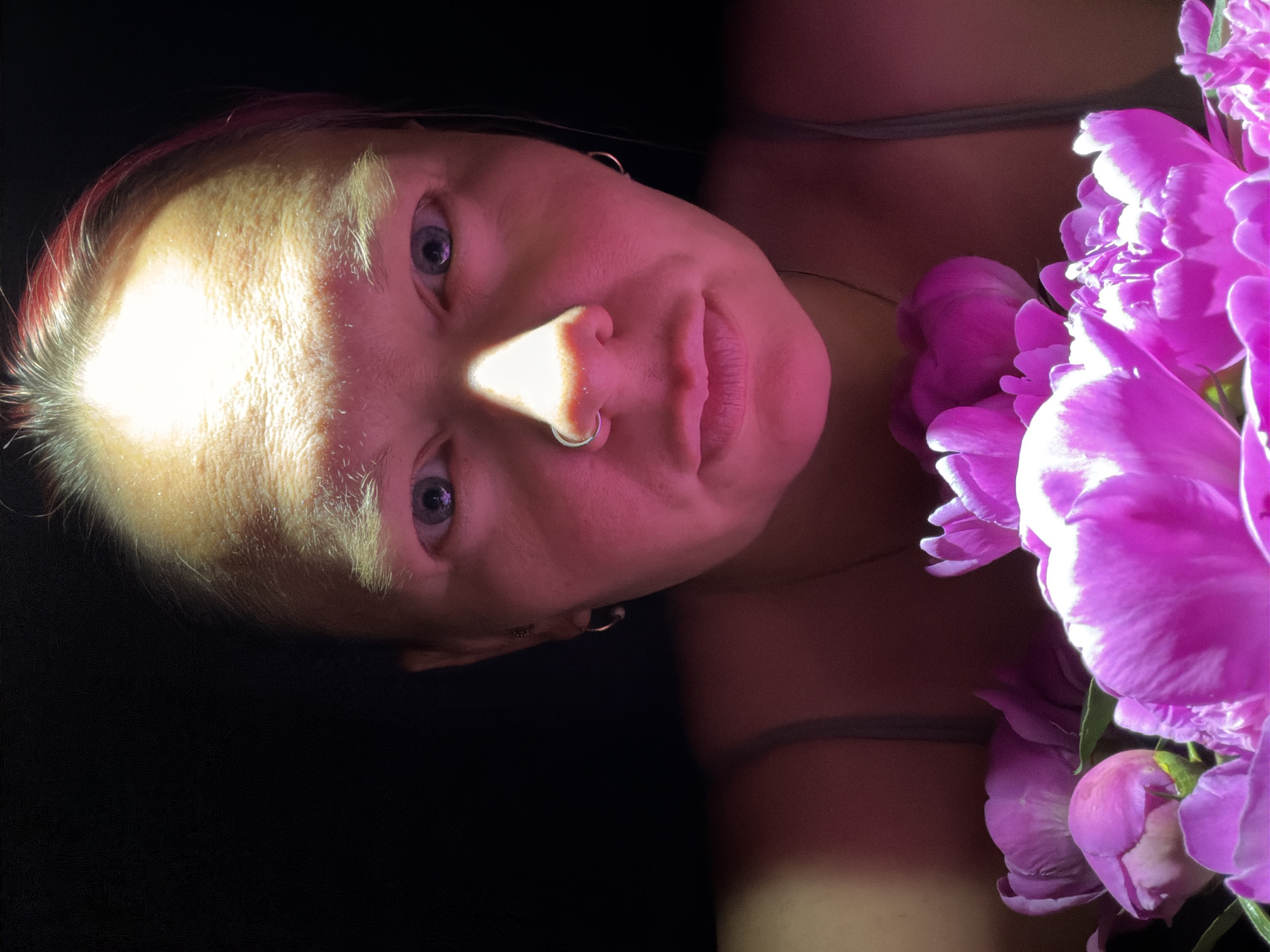 A close-up, low-angle studio shot depicts a person alongside pink peony blossoms. The individual has light skin, blonde hair with red streaks at the roots, blue eyes, and a nose ring. A strong light source casts a bright triangle on their forehead and nose. The person is wearing a light-colored top with thin straps. The background is predominantly dark, emphasizing the illuminated face and flowers. The location is listed as the city of Chaikovsky, Russia.