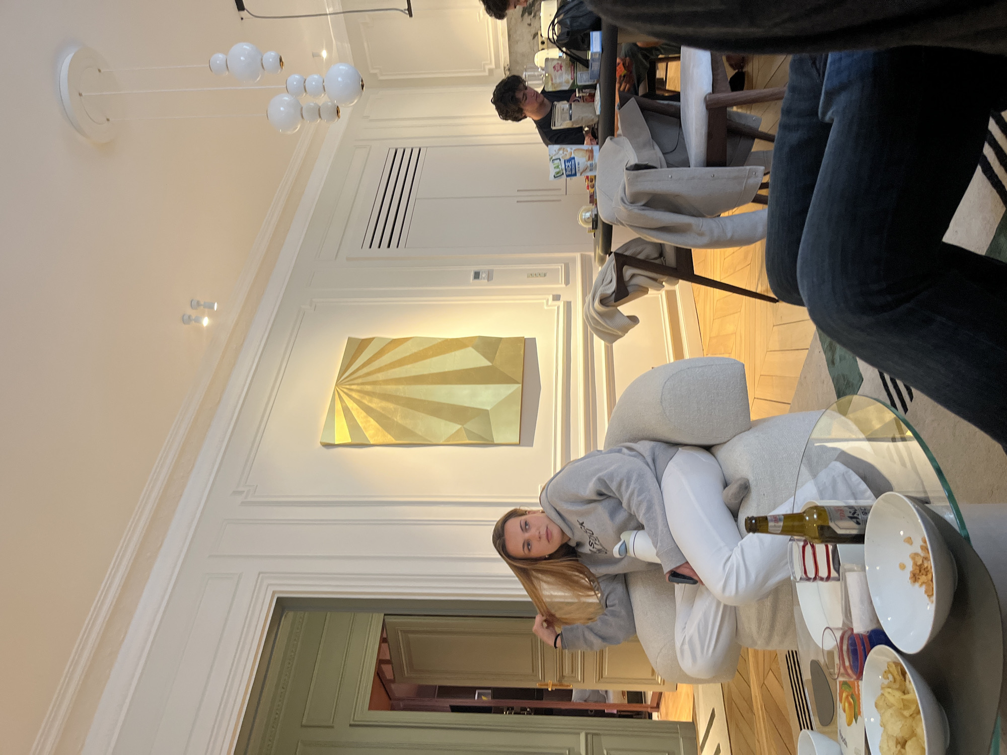 The image shows the interior of a stylish Parisian apartment or café in Paris, France. A young woman in a grey sweatshirt and white pants is reclining on a light-colored sofa. A glass coffee table in front of her holds snacks and a beer bottle. There are other individuals in the background, including someone at a table. The room features high ceilings, decorative moldings, modern lighting fixtures, and a golden abstract artwork. A door leads to another room with distinct green and beige accents.